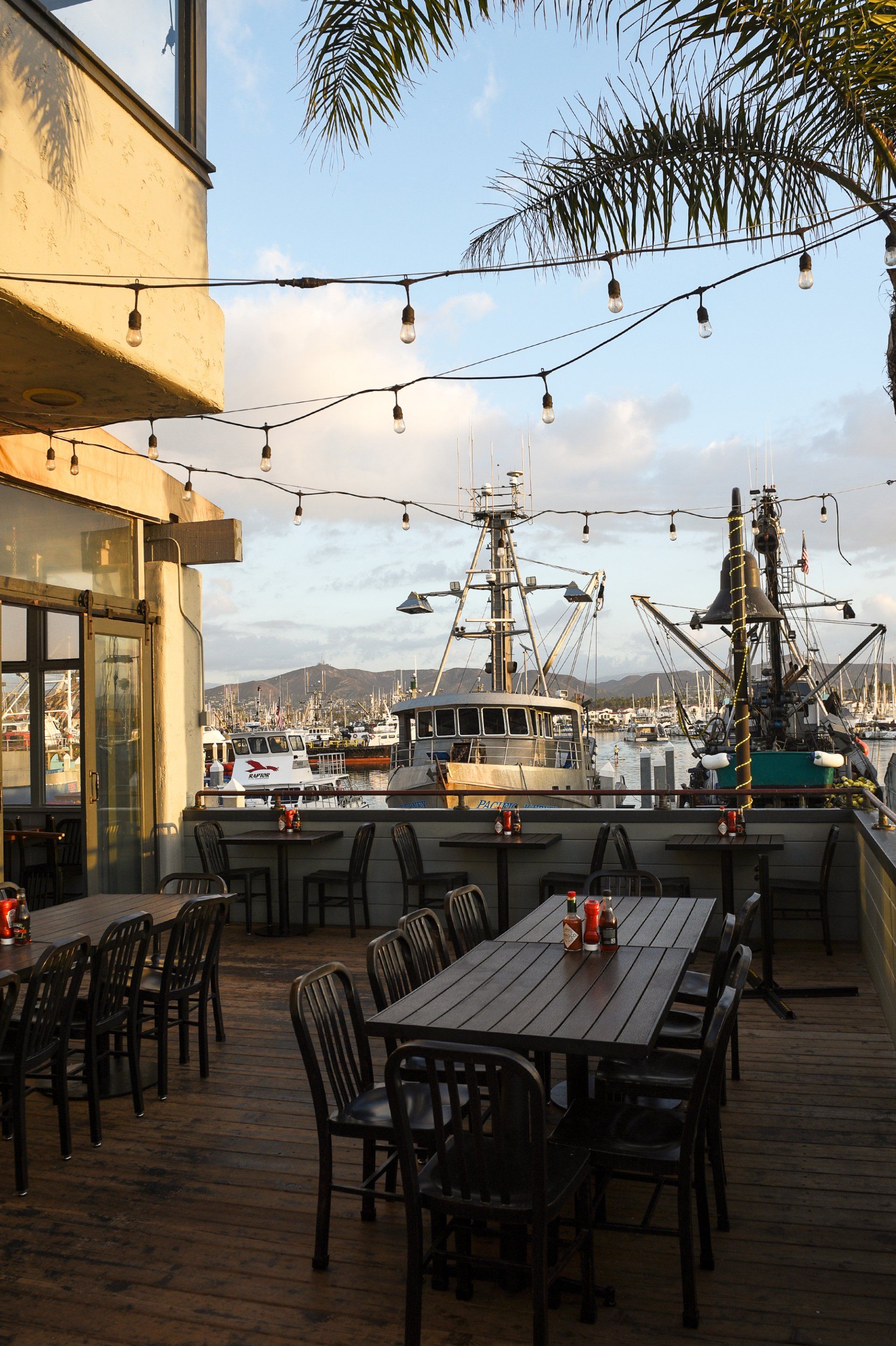 Outdoor restaurant patio overlooking a harbor with docked fishing boats under a string of hanging lights at sunset.