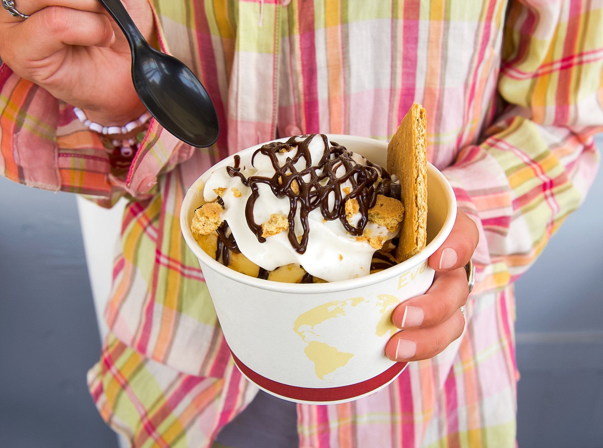 A person in a plaid shirt holds a white bowl of s'mores-style dessert topped with whipped cream and chocolate drizzle.