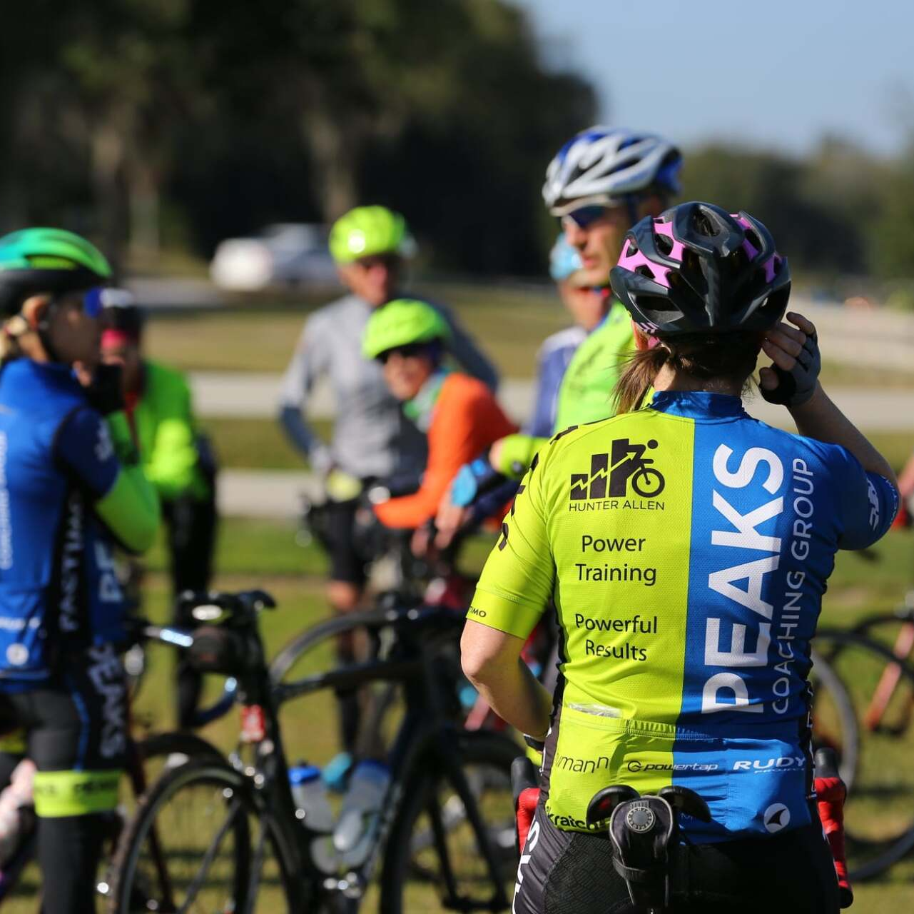 A woman wearing a peaks cycling group jersey stands in front of a group of cyclists.