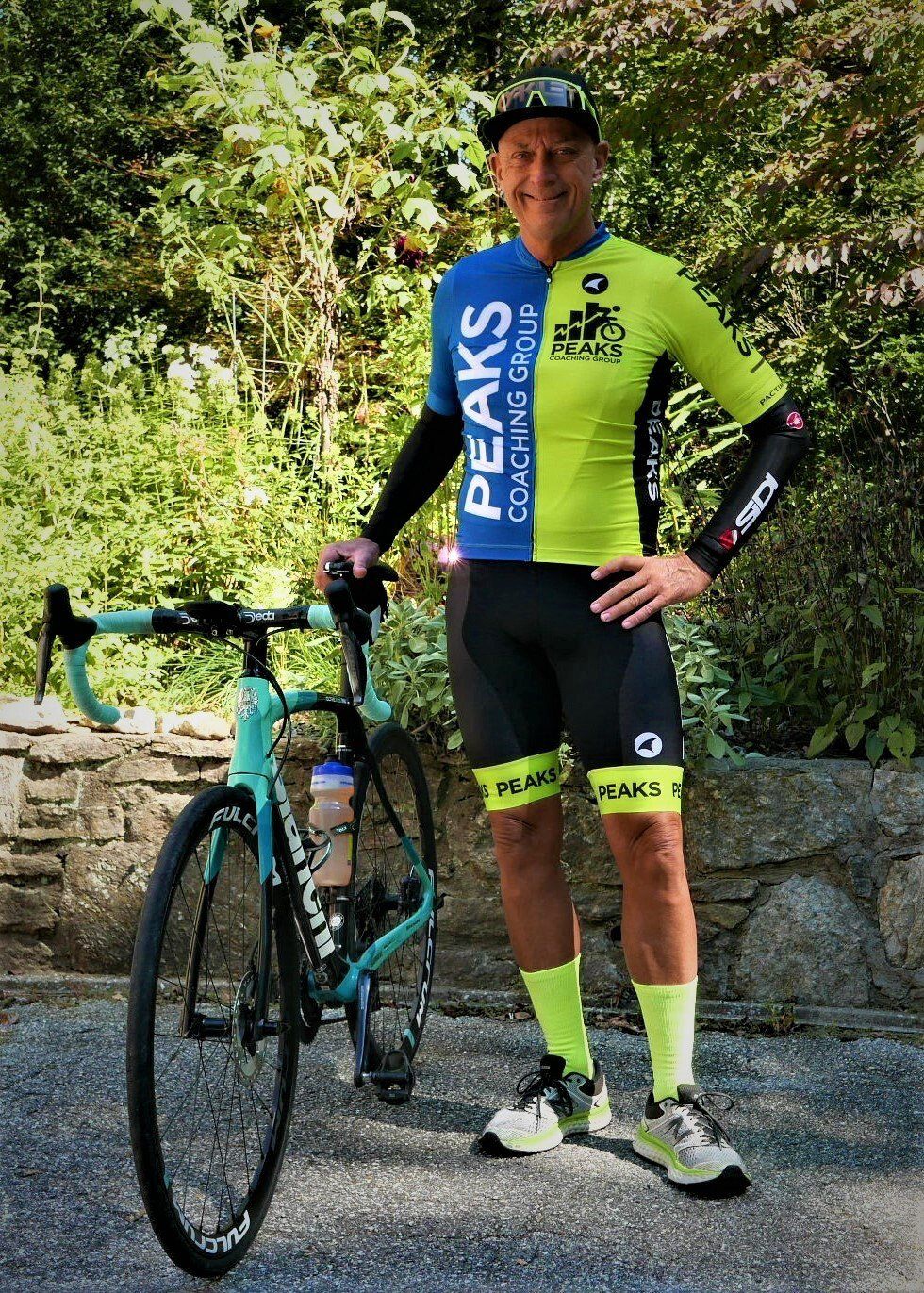 A man in a peaks cycling jersey is standing next to a bicycle.