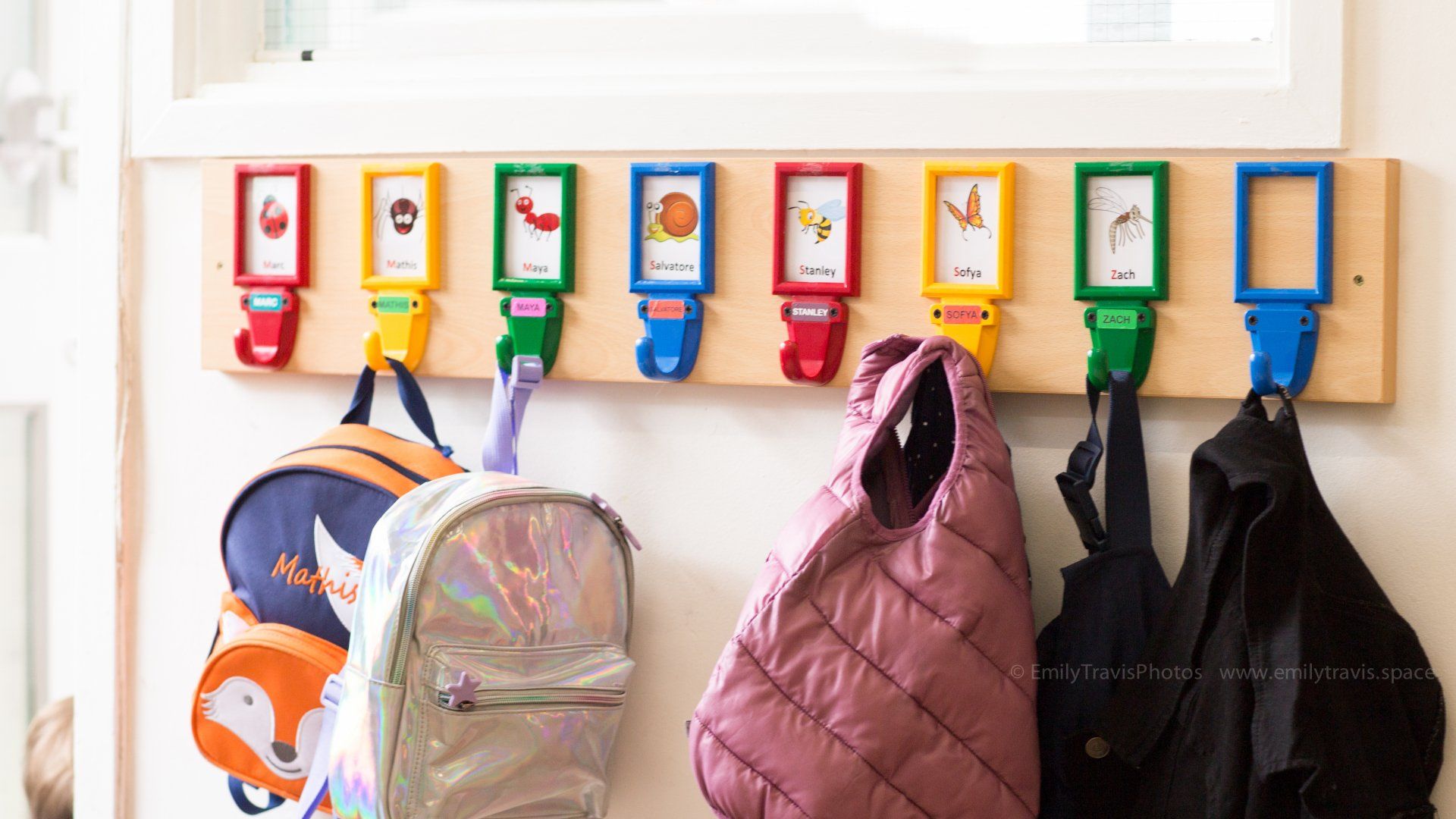 Colorful wall hooks holding three backpacks under a row of labeled cubbies in a classroom.