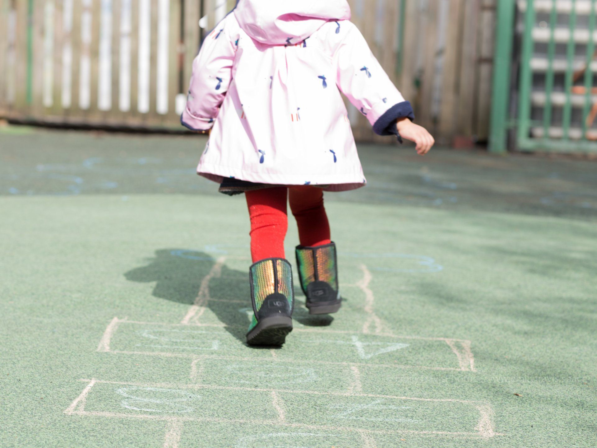 Child in a pink coat and red leggings stepping on hopscotch chalk on a playground.