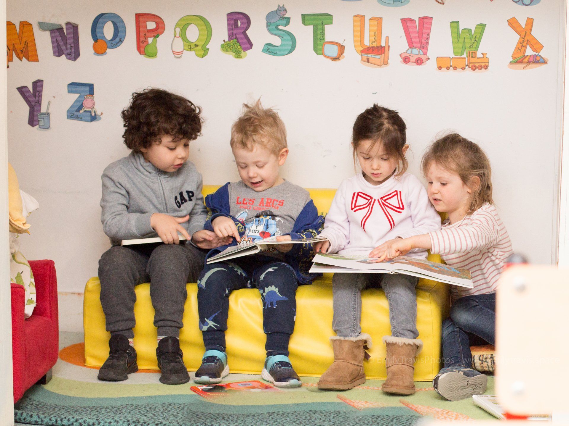 Children sitting on a yellow bench reading books in a colorful classroom with alphabet letters on the wall