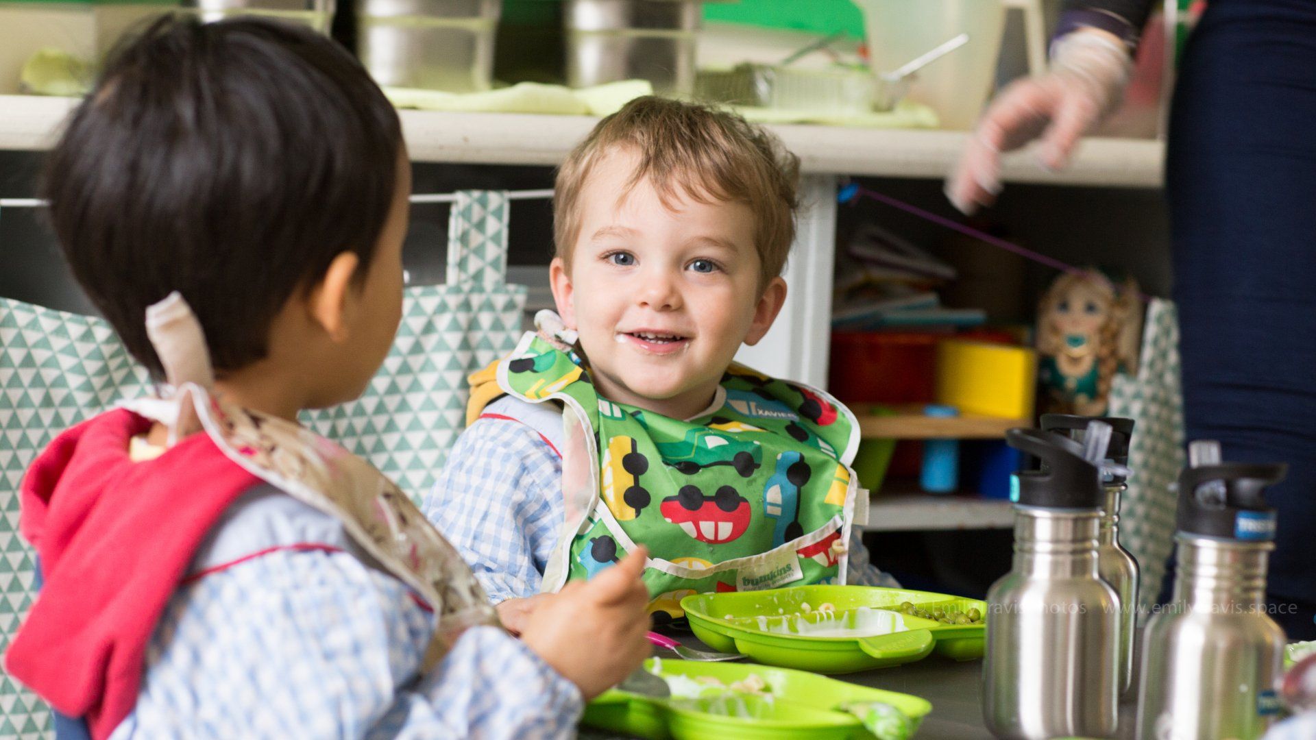 Children eating at a table in a daycare, with one smiling toward the camera and green plates visible