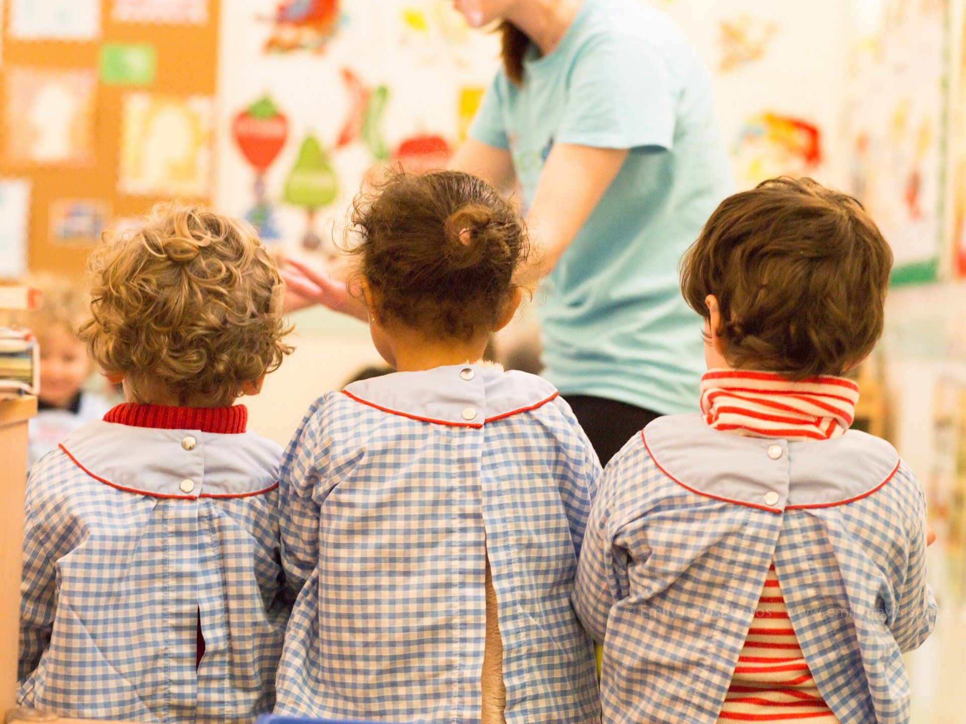 Three children in checkered smocks sit facing a teacher in a colorful classroom