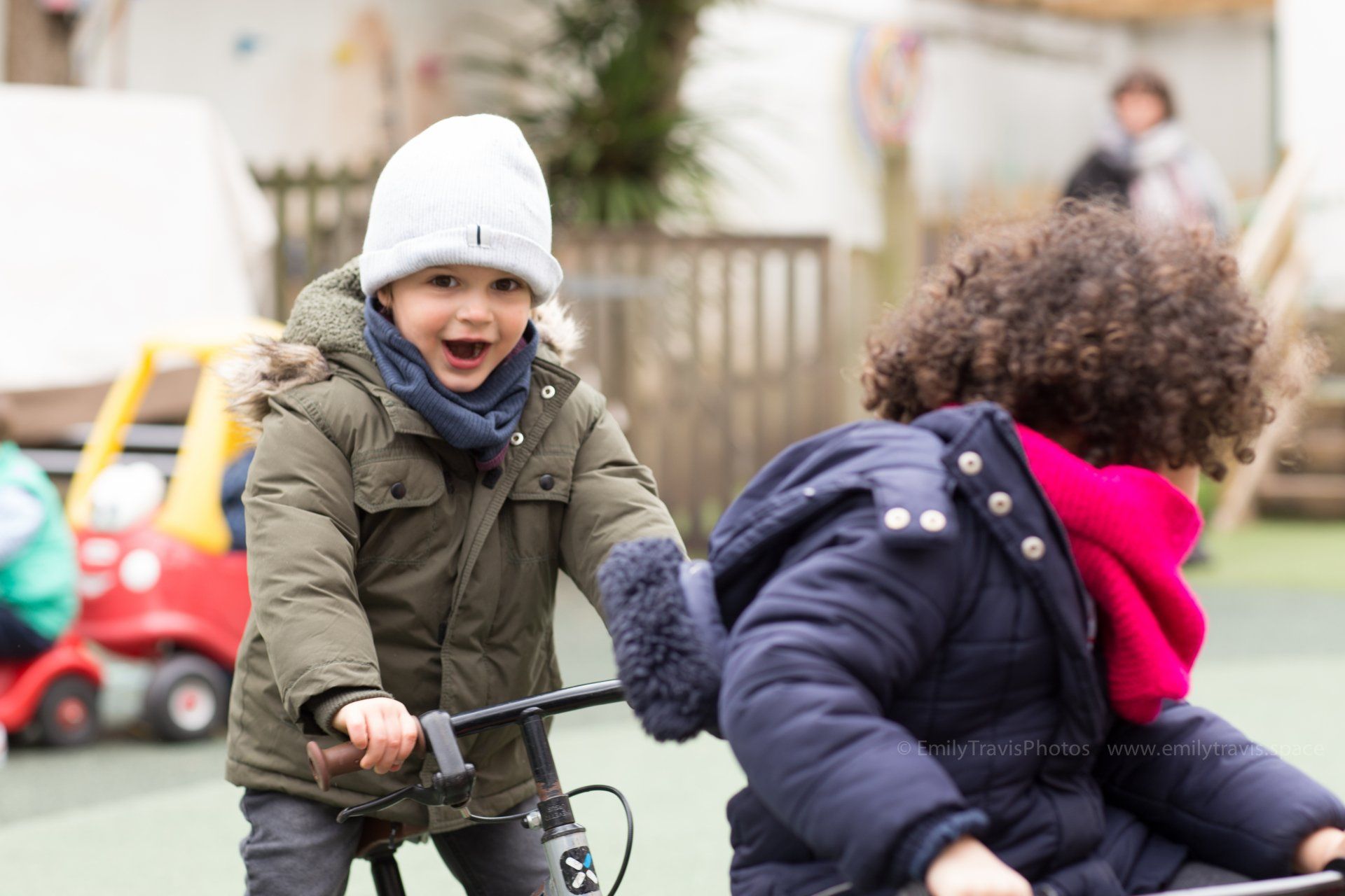 Two children riding balance bikes outdoors, one smiling in a white hat and green coat