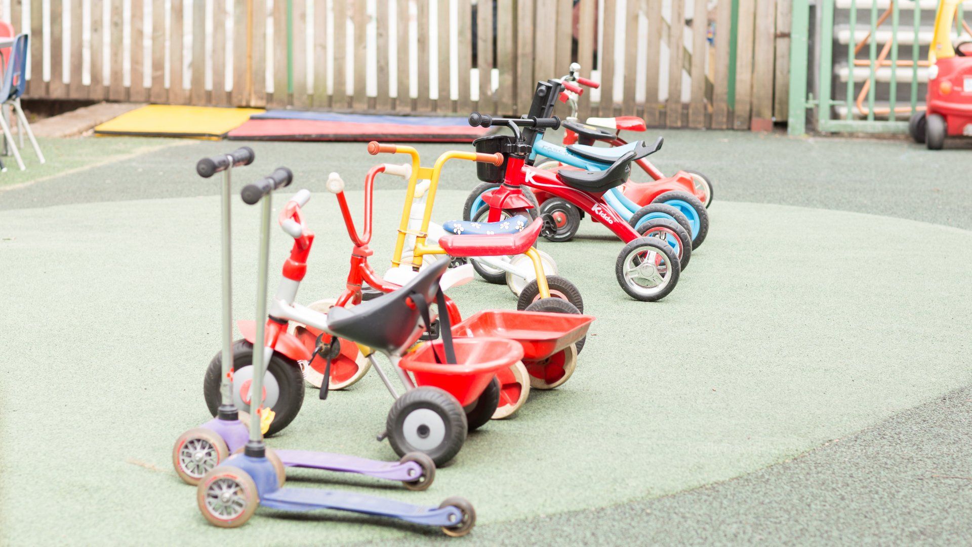 Children’s scooters and ride-on toys parked on a playground surface near a wooden fence