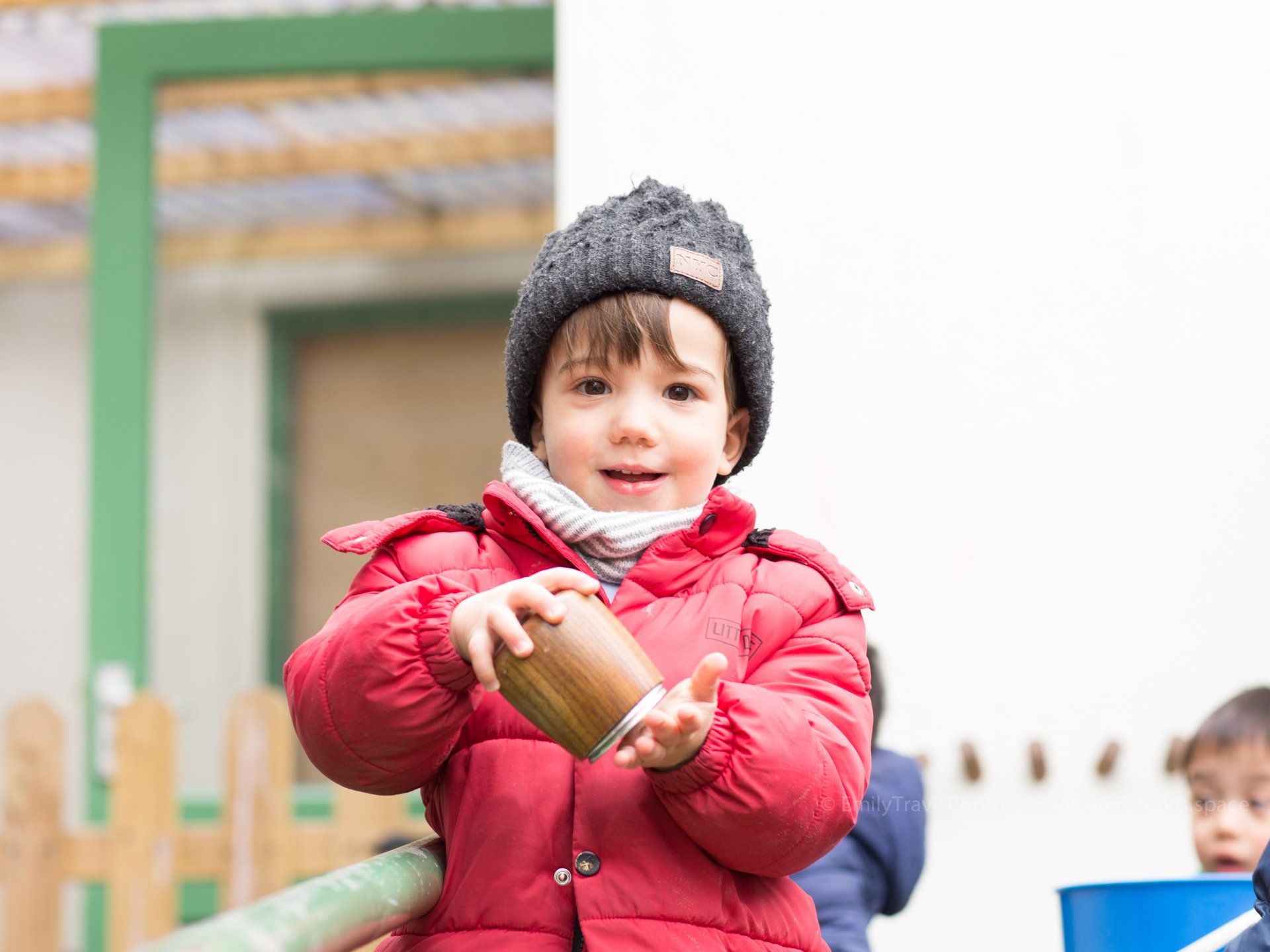 Child in a red coat and knit hat holding a small cup outdoors, with a playground fence in the background