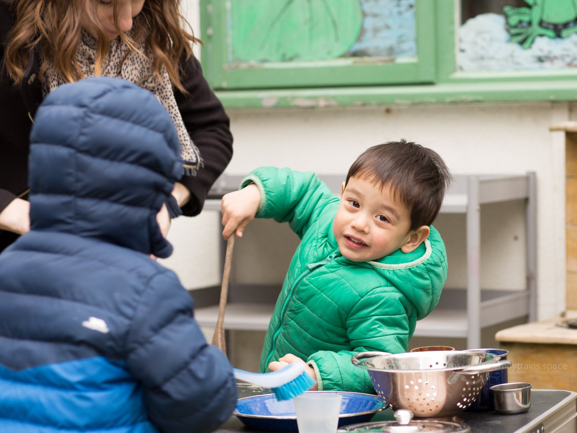 Children playing with kitchen utensils at a table, one in a green jacket smiling and stirring a pot