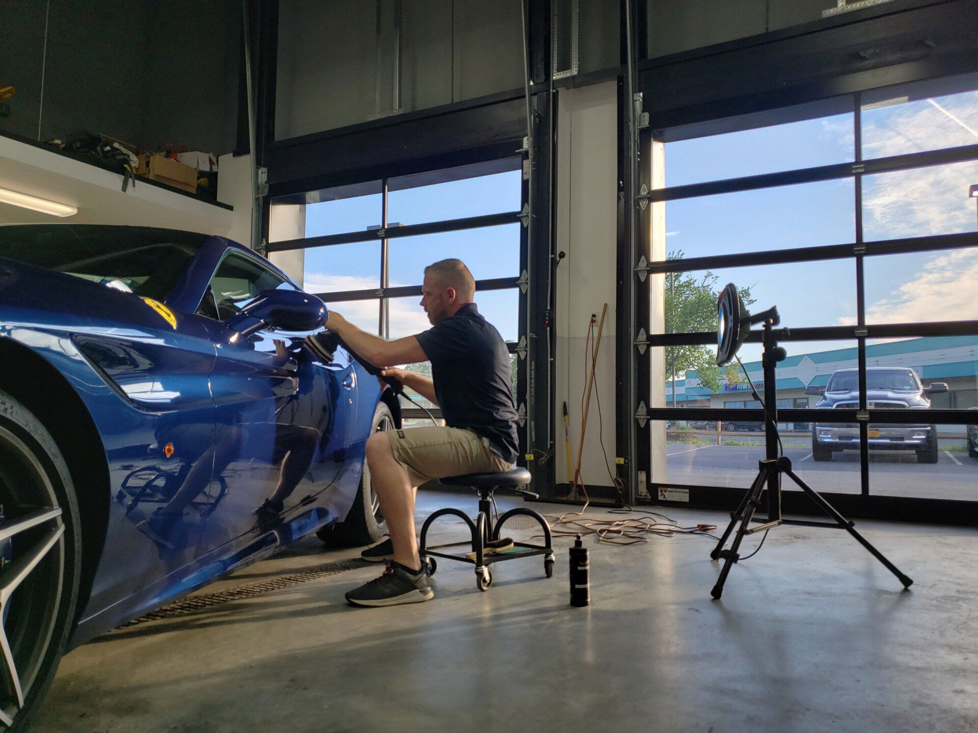 A man is sitting on a stool in front of a blue car in a garage.