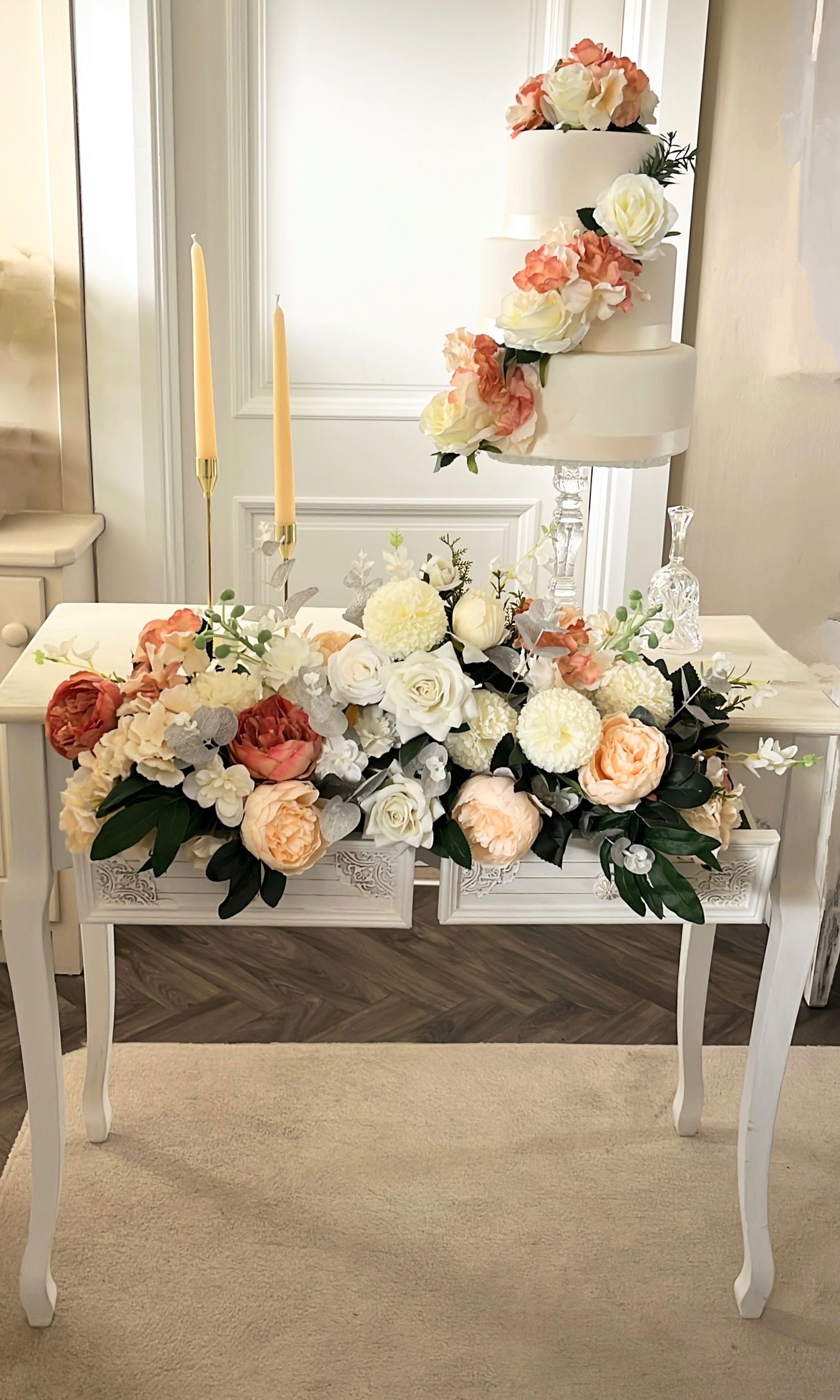 White floral wedding table display with peach and cream arrangements, candles, and a tiered flower stand