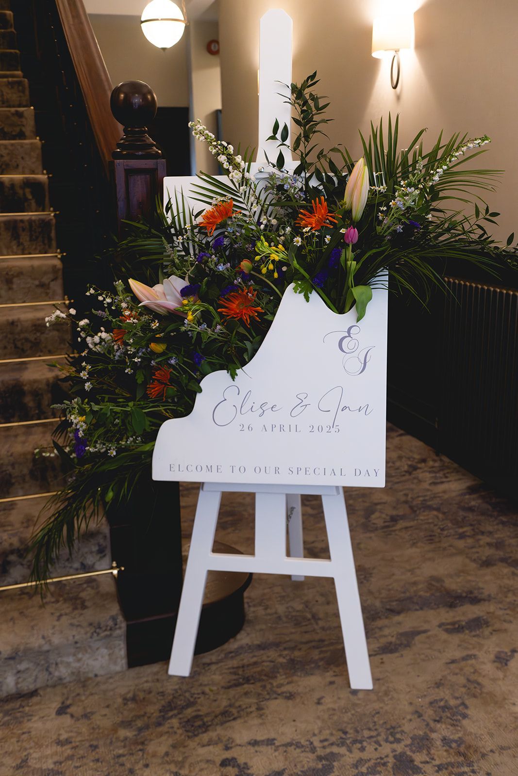 Floral arrangement on a white easel at an indoor staircase entrance, with greenery and colorful flowers.