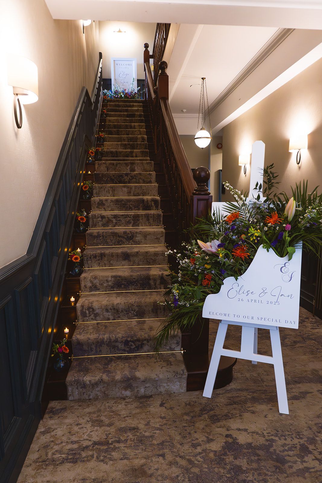 Staircase in a decorated hallway with flowers and a white sign on an easel at the base.