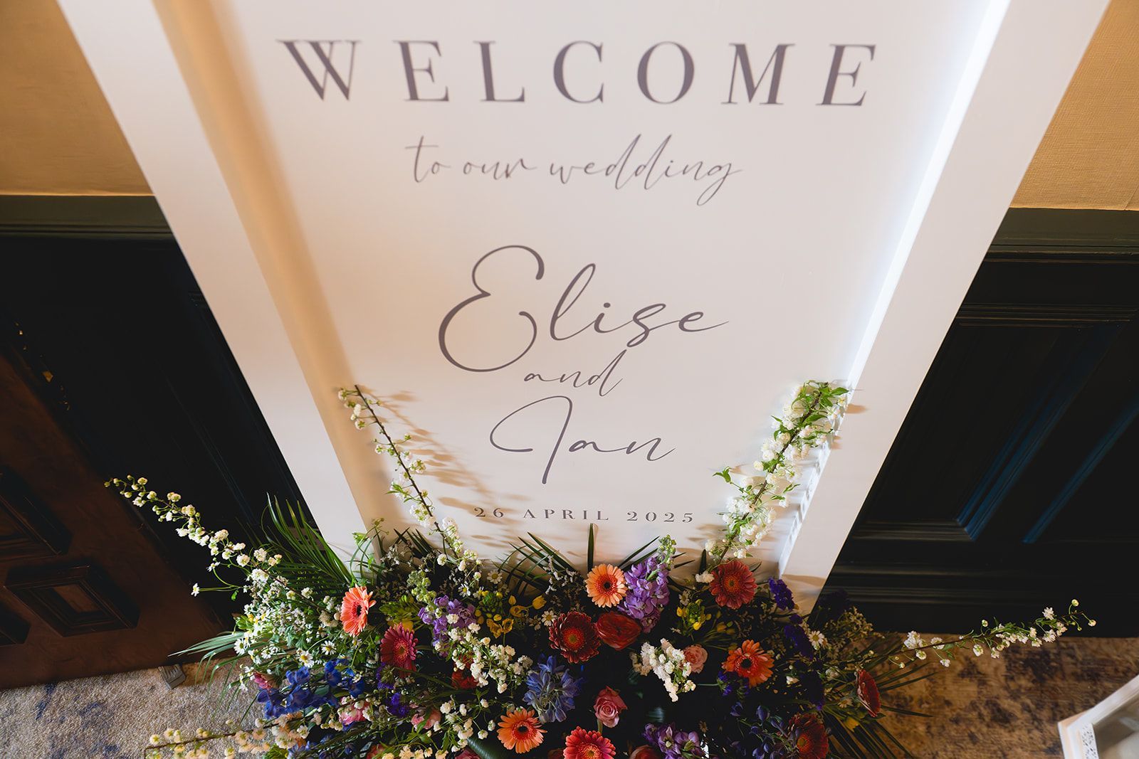 Wedding welcome sign with floral arrangement at the base, in white, gold, and purple tones
