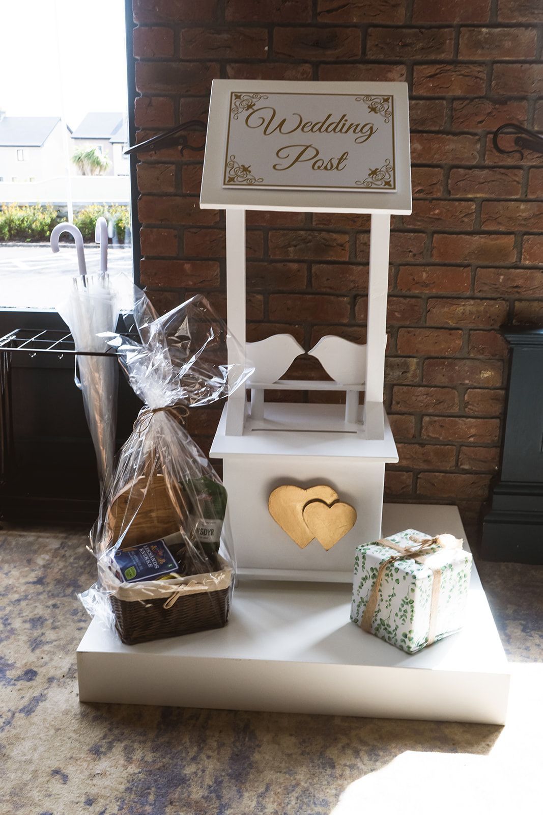 Decorative white wedding card box with heart cutout, baskets, and a “Wishing you…” sign on a brick wall backdrop