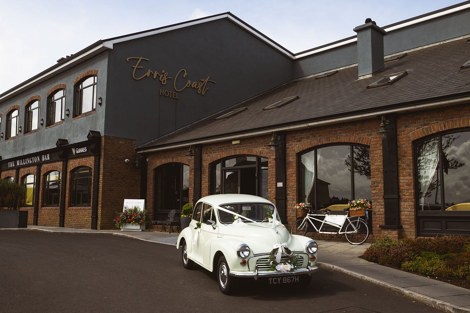 White vintage car parked outside a brick hotel or restaurant with large windows and signage.