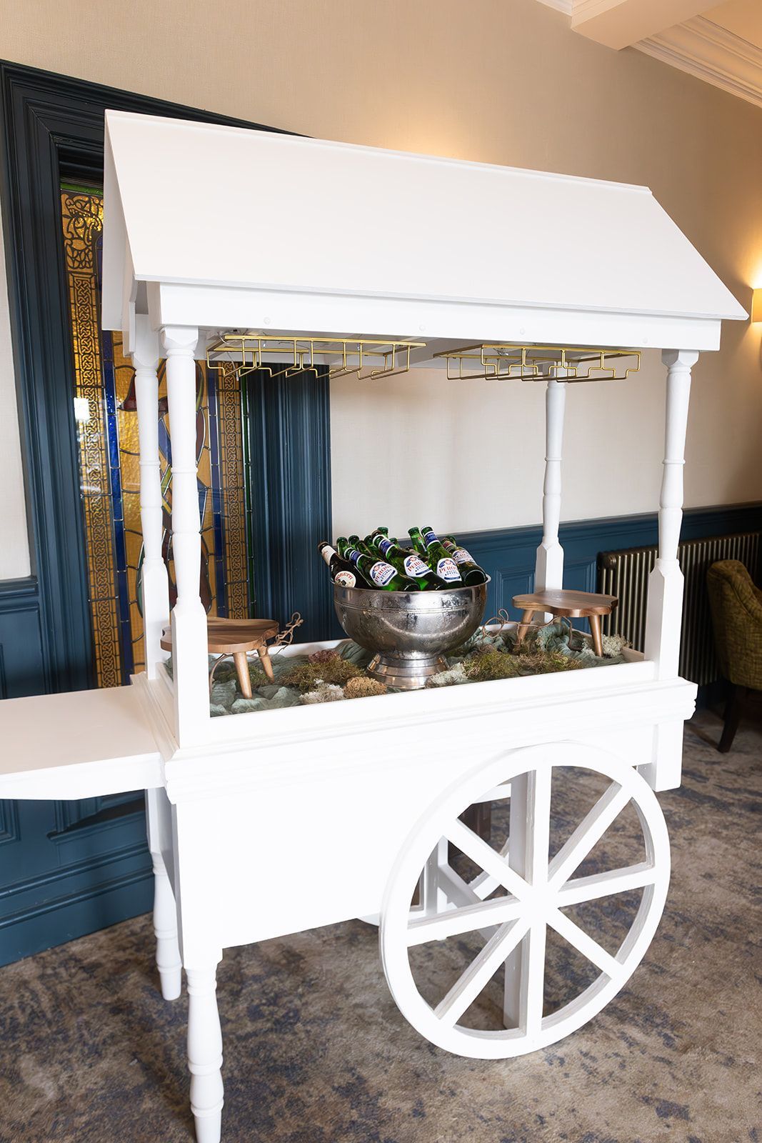 White decorative cart with awning and large wheel, holding flowers and glassware indoors