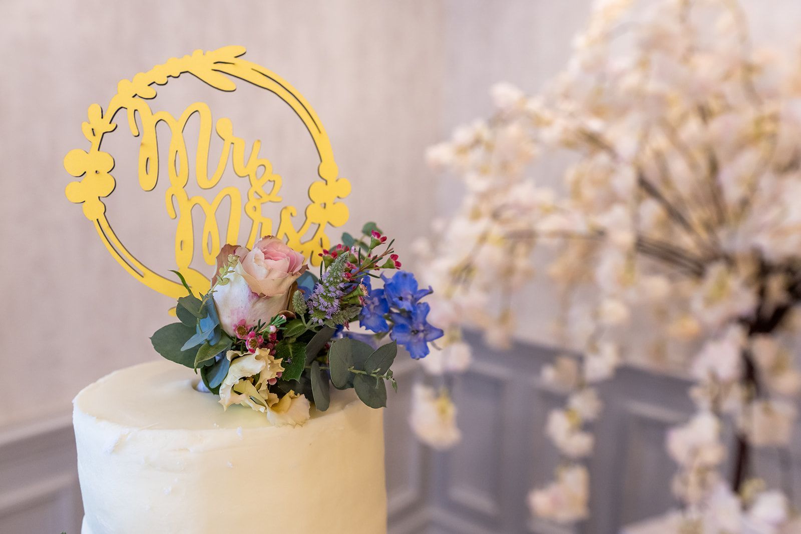 White cake topped with floral arrangement and gold “Mr & Mrs” topper at a wedding reception