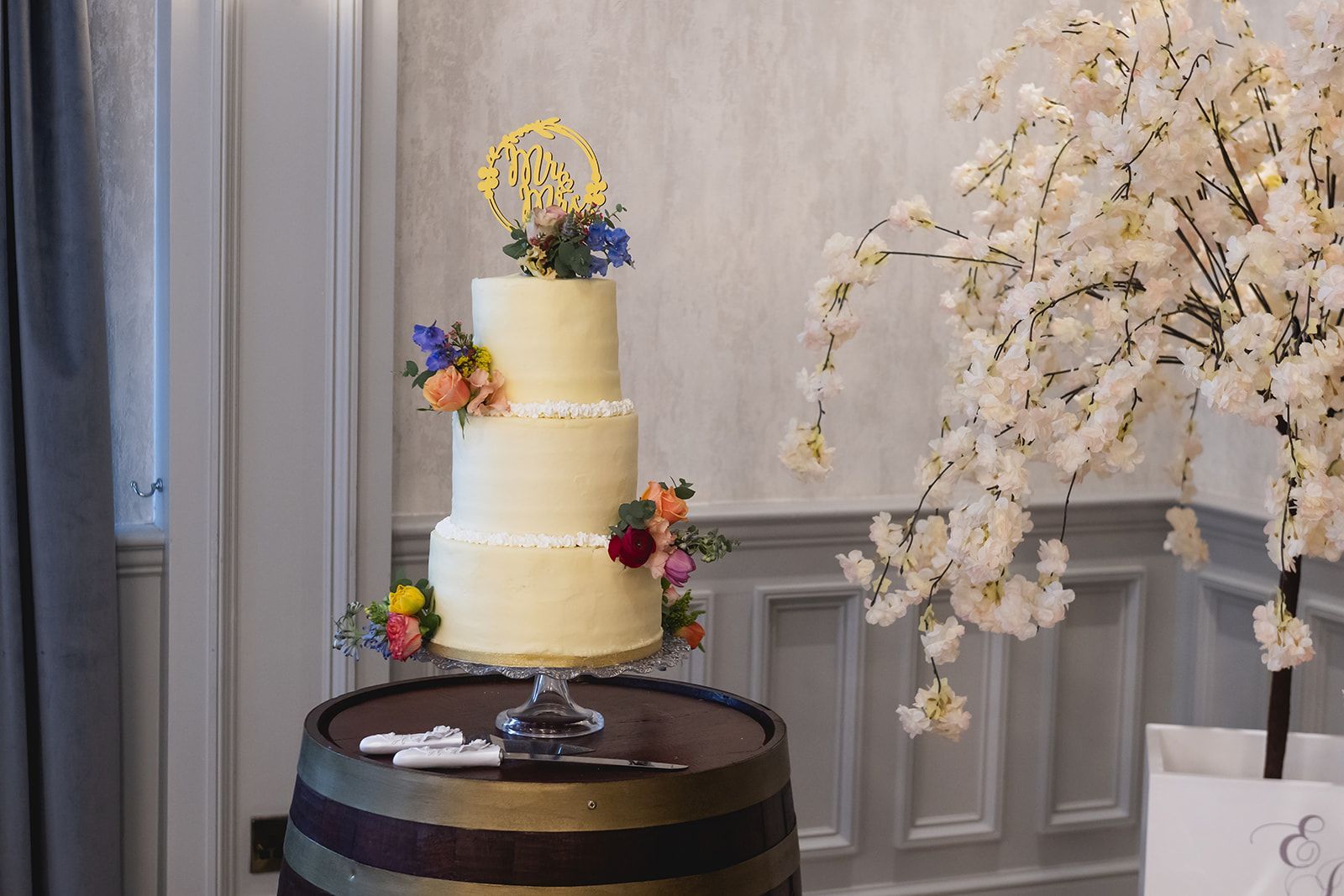 Two-tier cream wedding cake with colorful flowers on a barrel table beside white cherry blossoms.