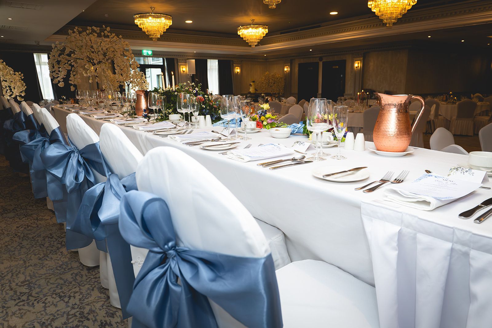 Elegant banquet hall with long white tablecloths, blue chair sashes, and chandeliers overhead