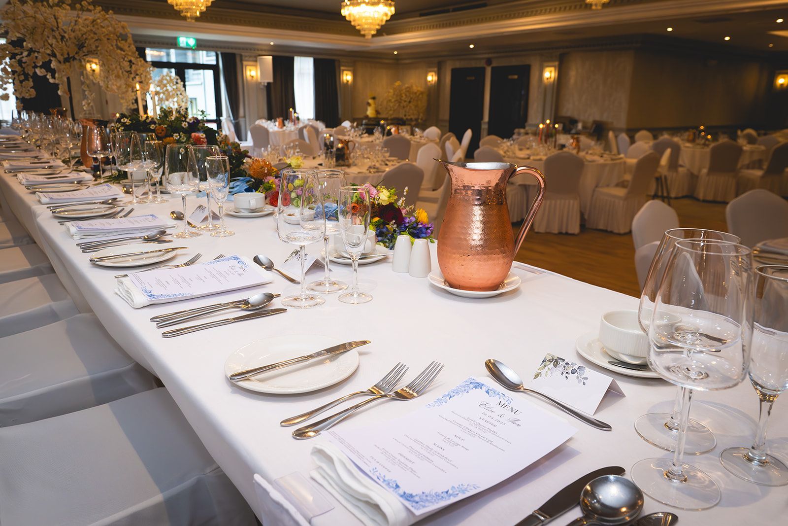 Banquet table set with white linens, place settings, candles, and floral centerpieces in a dimly lit hall
