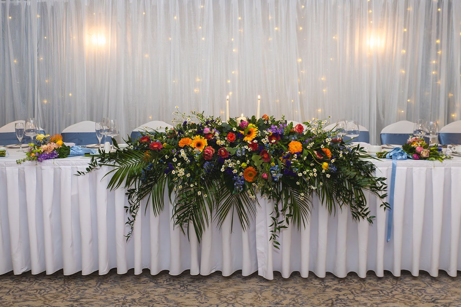 Decorated banquet table with white tablecloth, colorful floral centerpiece, and string lights backdrop