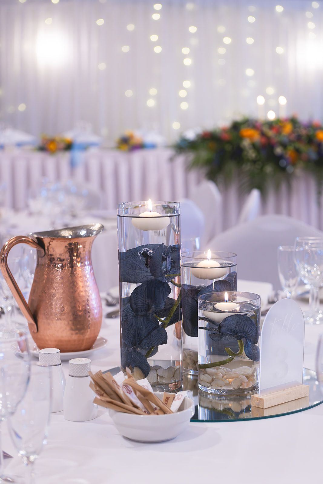 Elegant table centerpiece with copper pitcher, candles, blue stones, and fairy lights in a softly lit reception setting