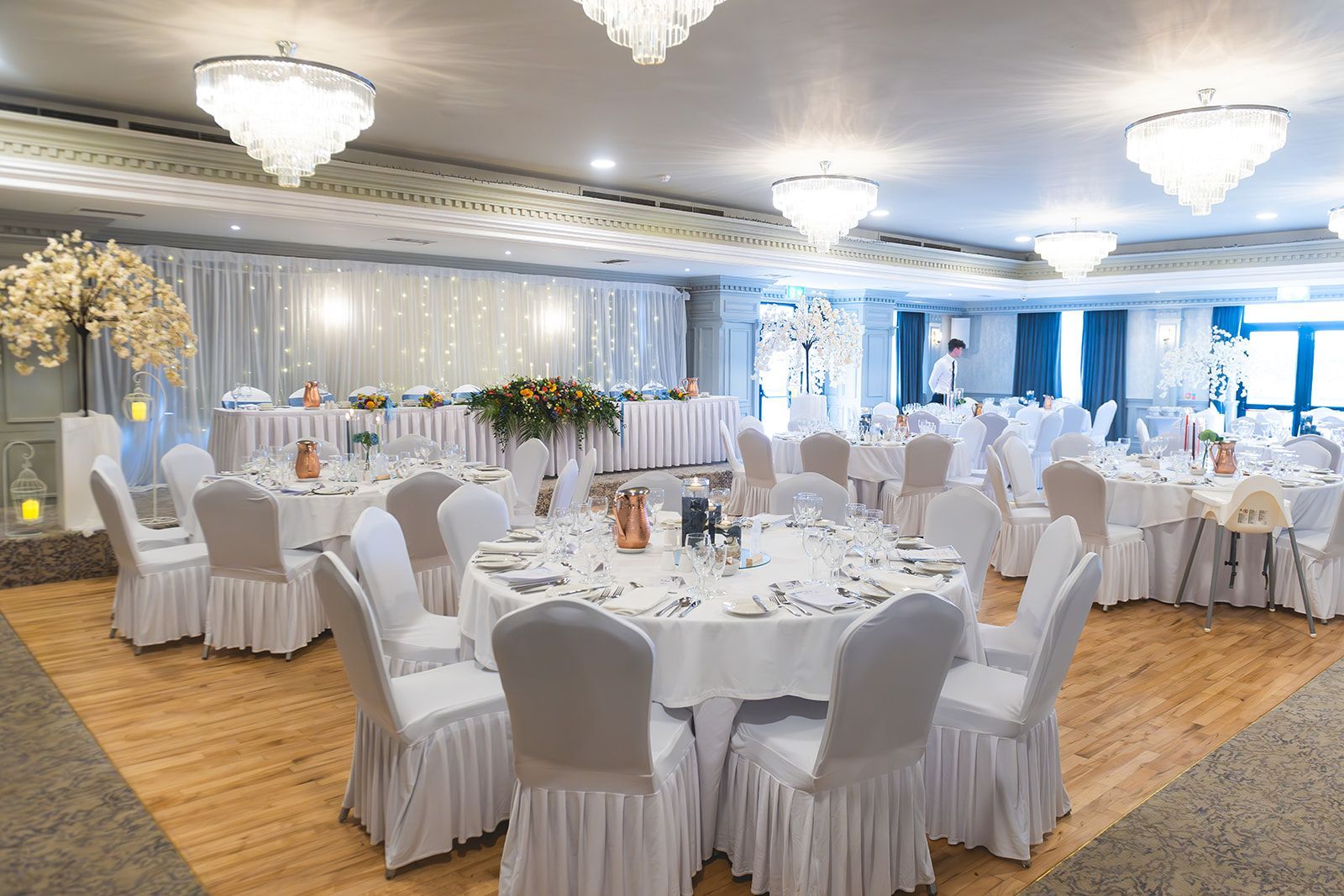 Elegant banquet hall with round white-clothed tables, white chairs, chandeliers, and blue lighting