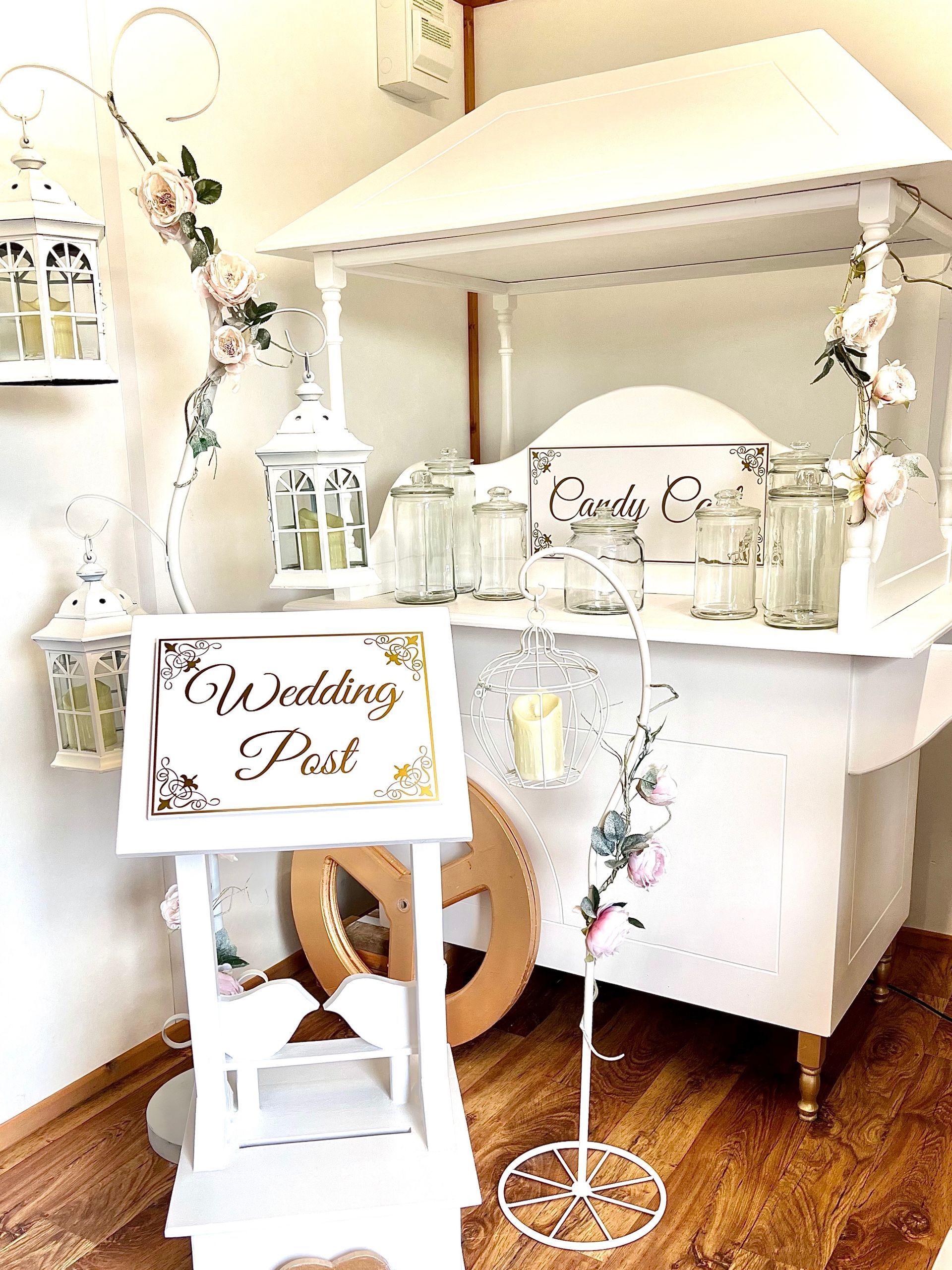 White wedding dessert and drink station with “Our Wedding” sign, lanterns, flowers, and wooden floor