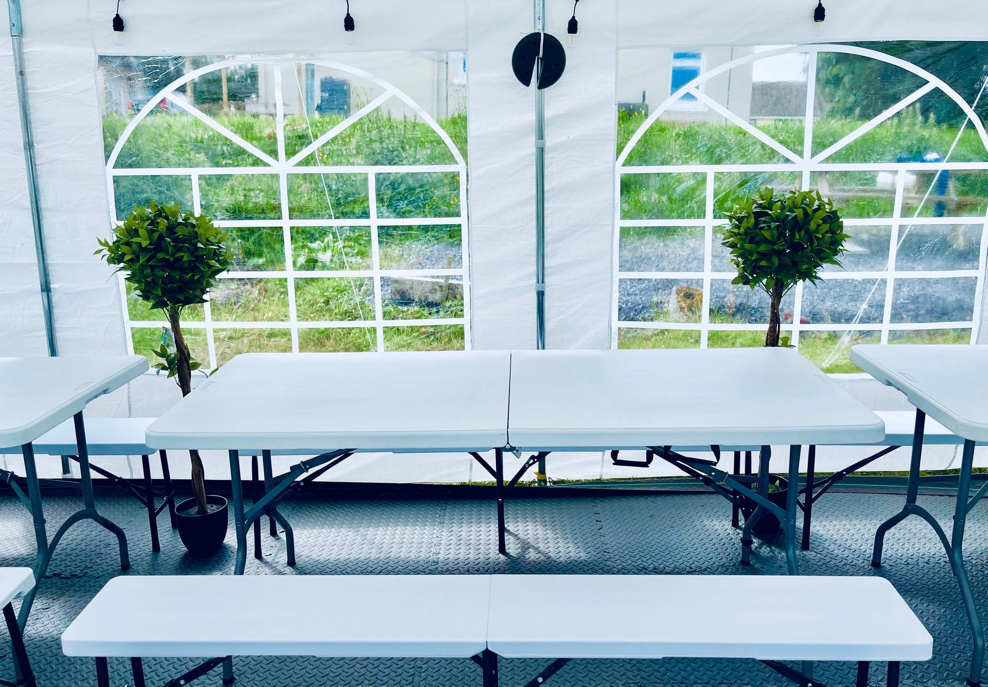 Empty white banquet tables in a bright tent with arched windows and green trees outside