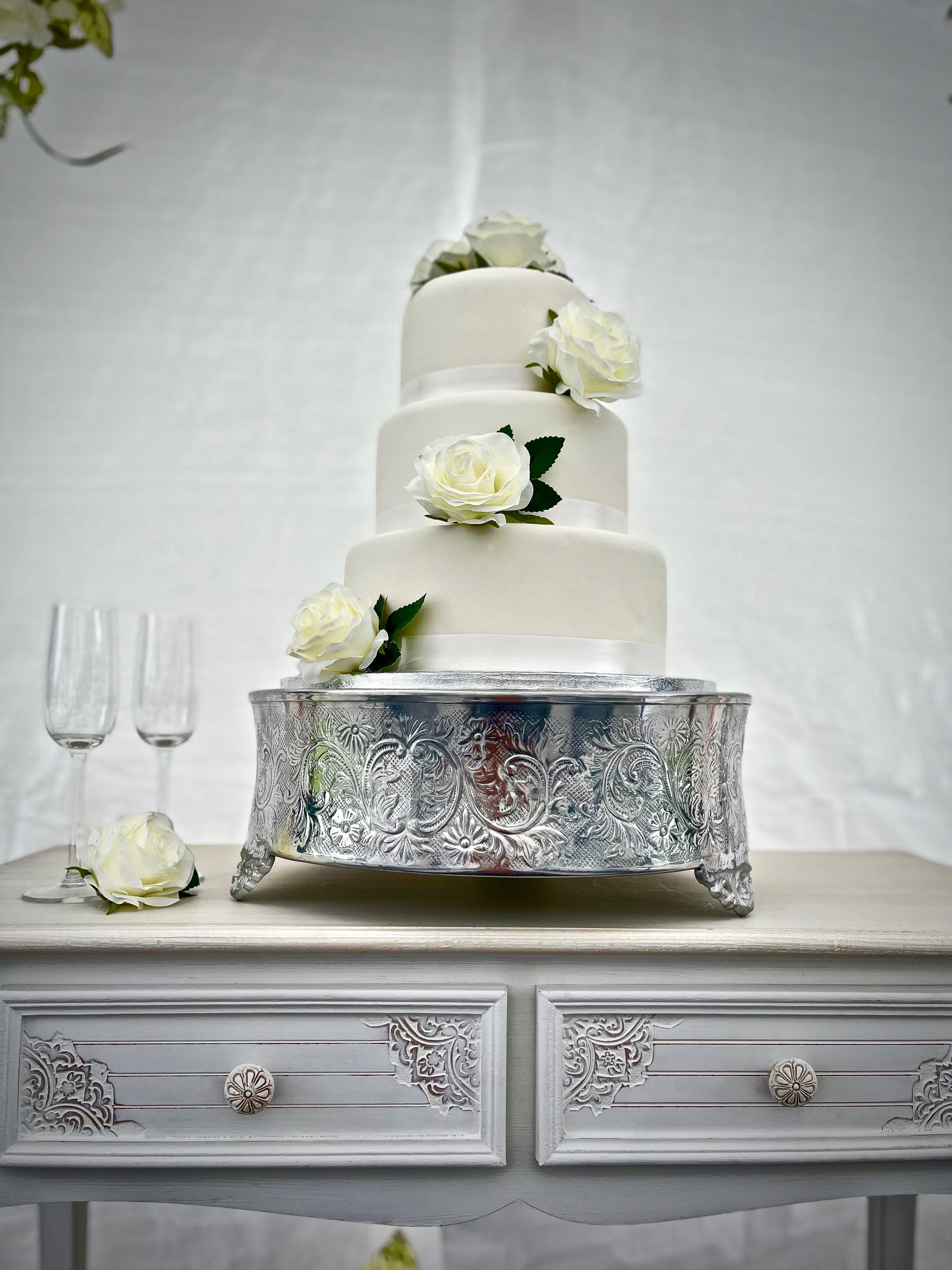 Three-tier white wedding cake with white flowers on a silver table, beside two empty champagne flutes.