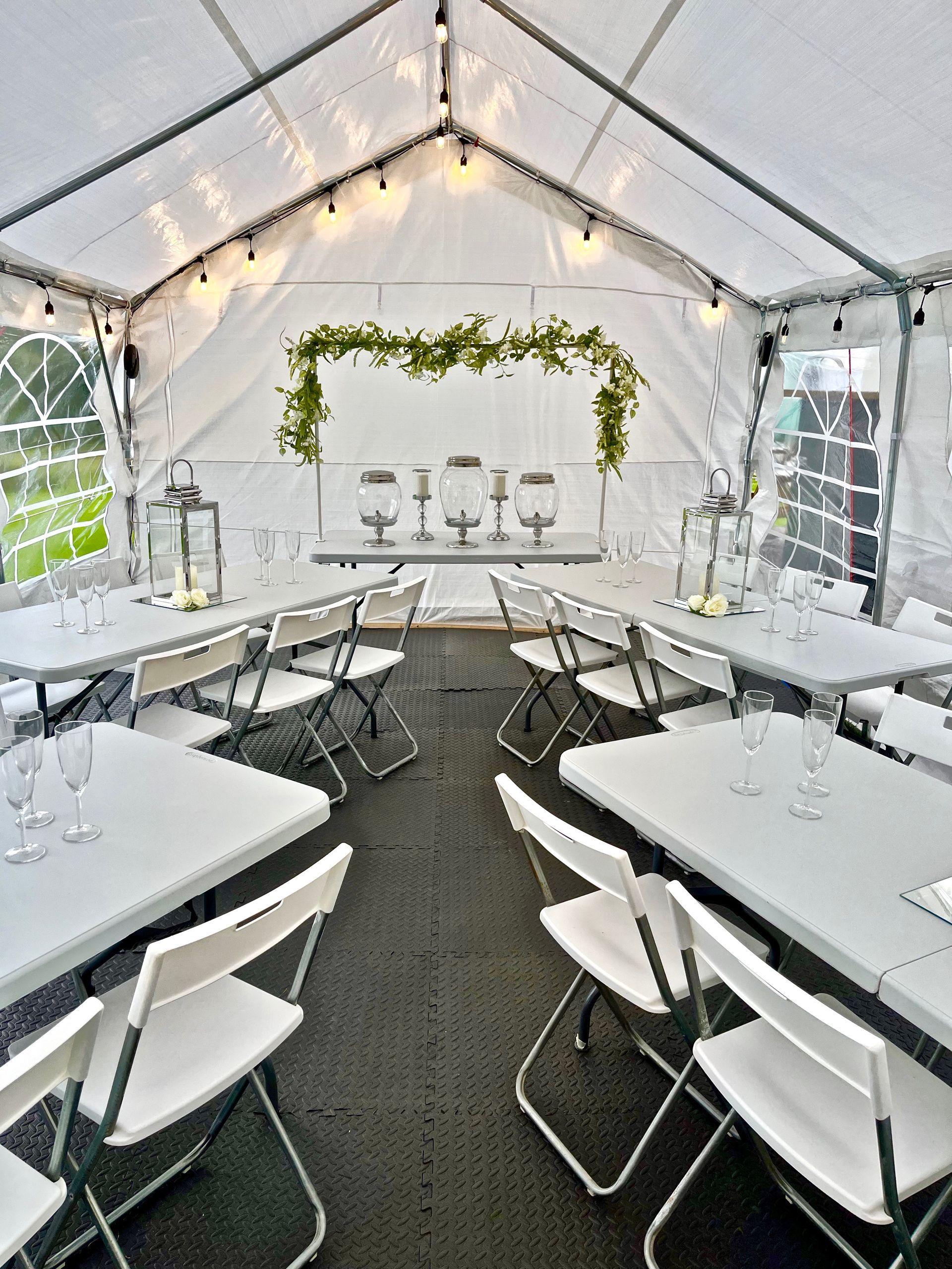 White tent reception setup with string lights, greenery backdrop, and round tables with white chairs
