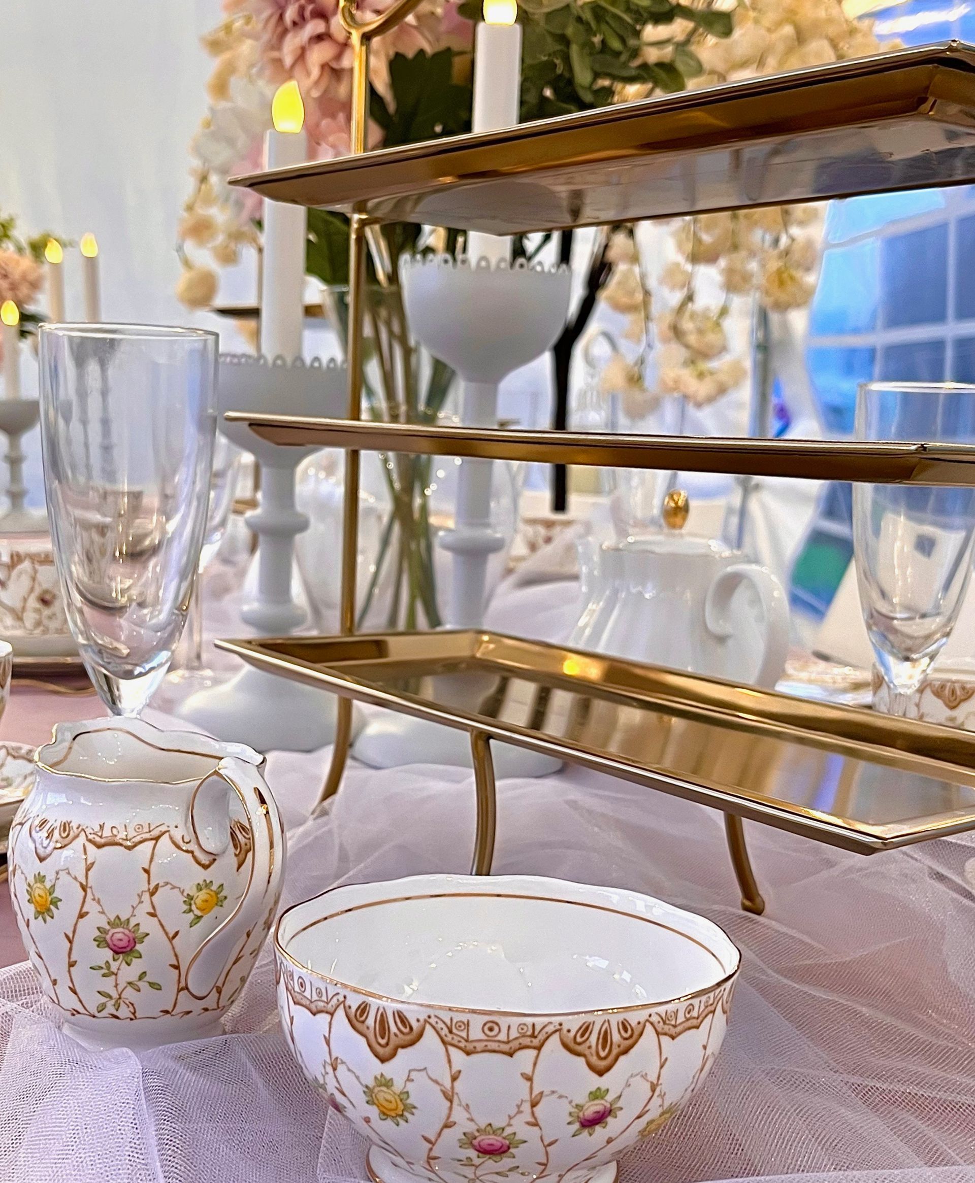 Elegant tea table with gold-trimmed porcelain cups and a tiered dessert stand in a soft pink setting