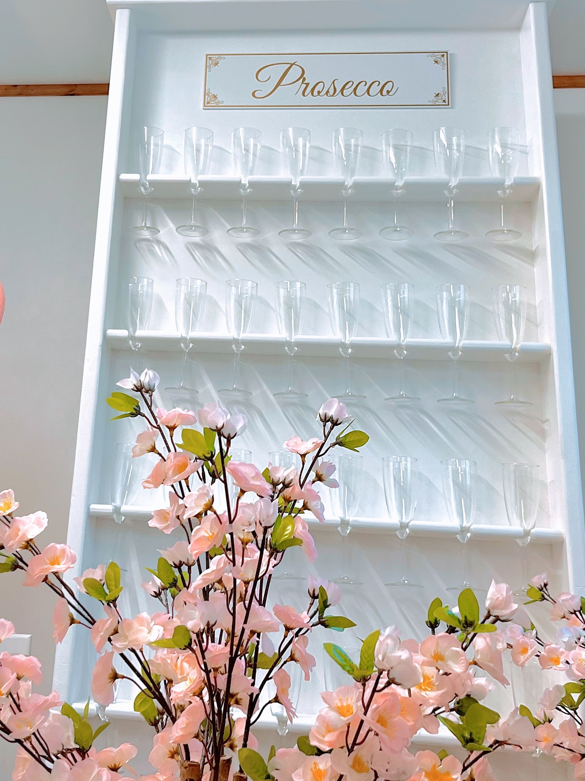 White shelf with a gold “Princess” sign and rows of glassware above pink blossoms