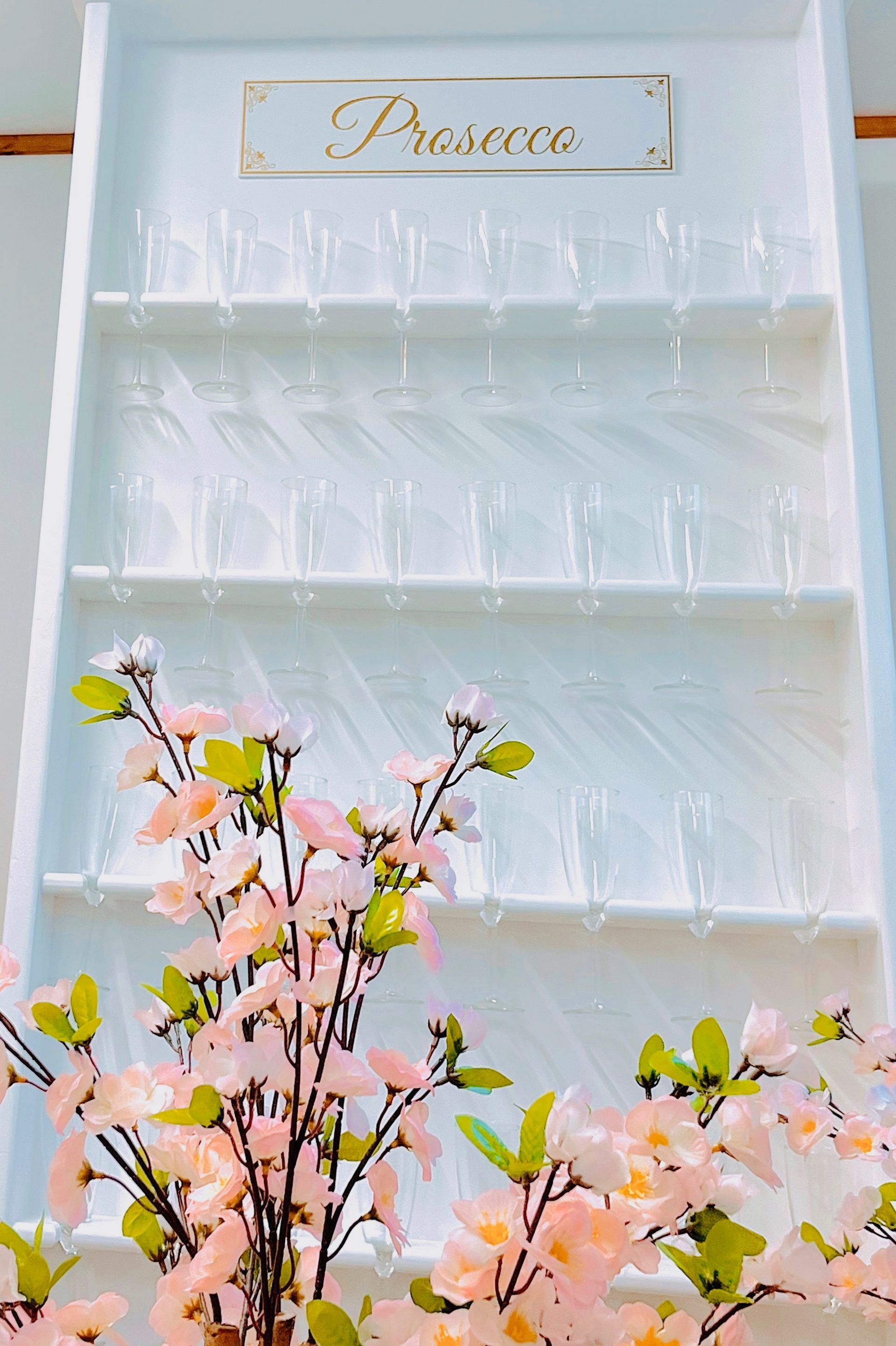 White decorative shelf with glassware and pink flowers beneath a gold “Pizzeria” sign.