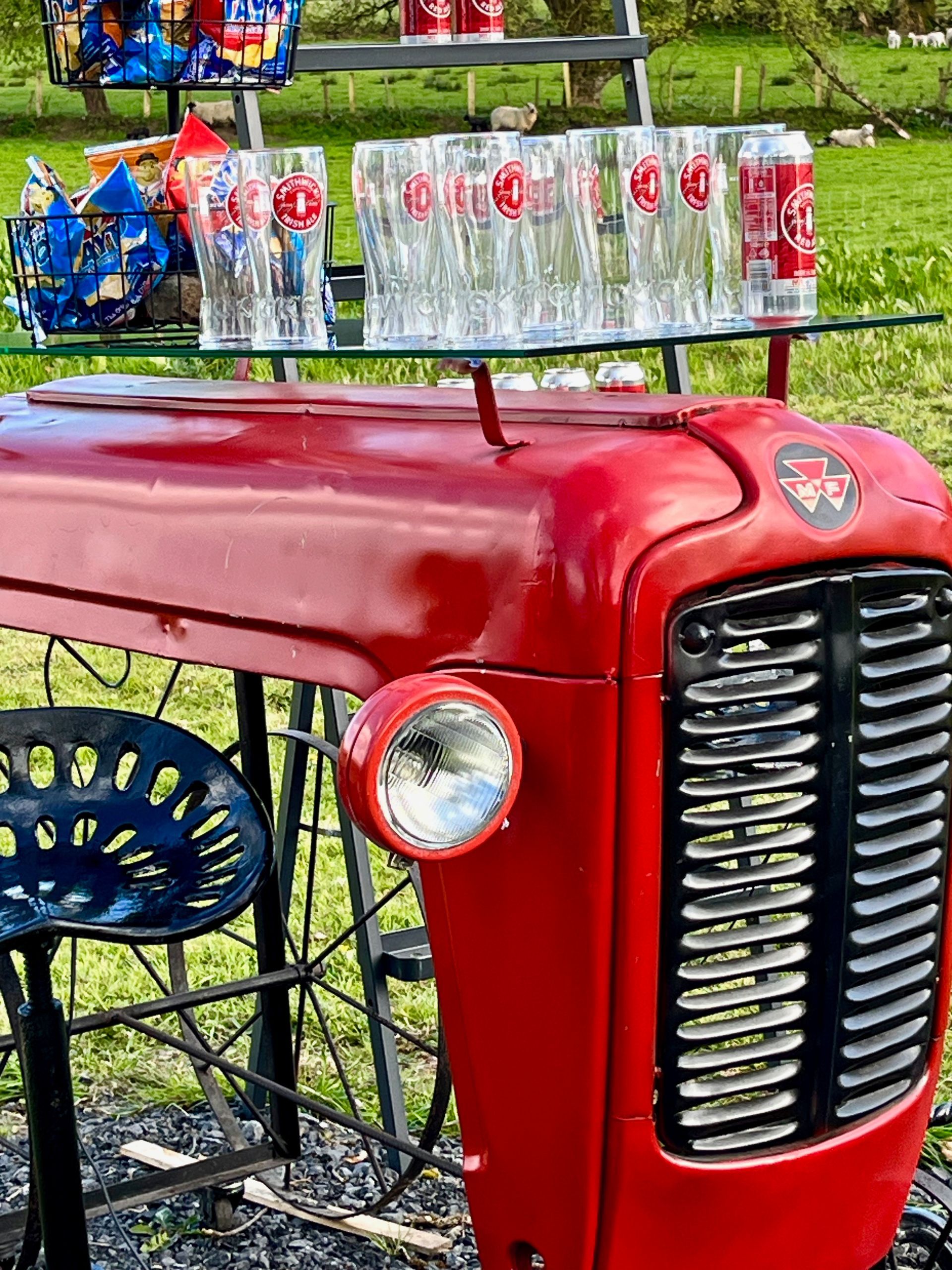 Red vintage tractor front with chrome grille and a row of cans on top, outdoors on grass.