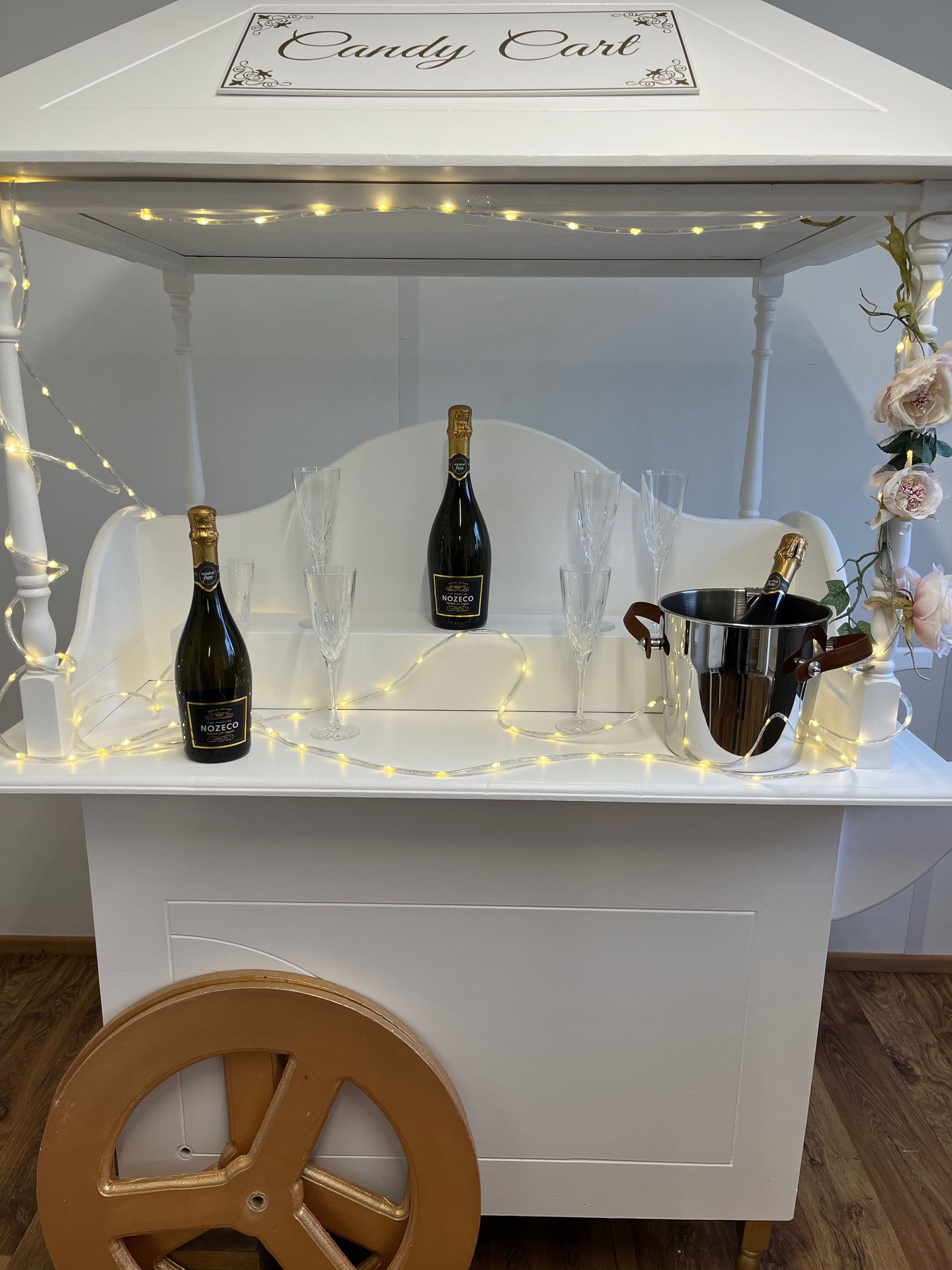 Decorative champagne display with three bottles, fairy lights, and a white cart on a wooden wheel in a white room