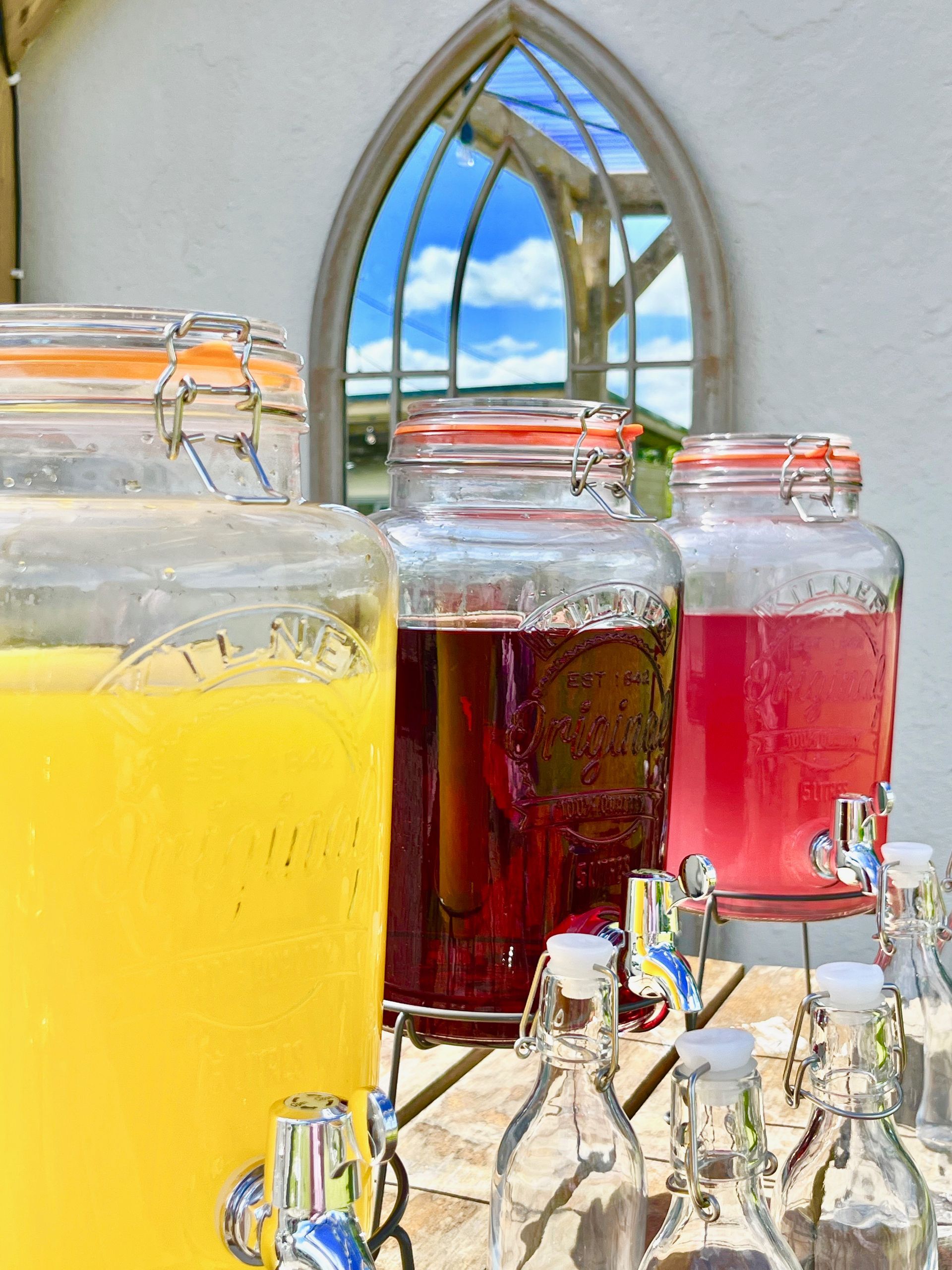 Three colorful beverage dispensers with drinks on a table, with a bright window in the background