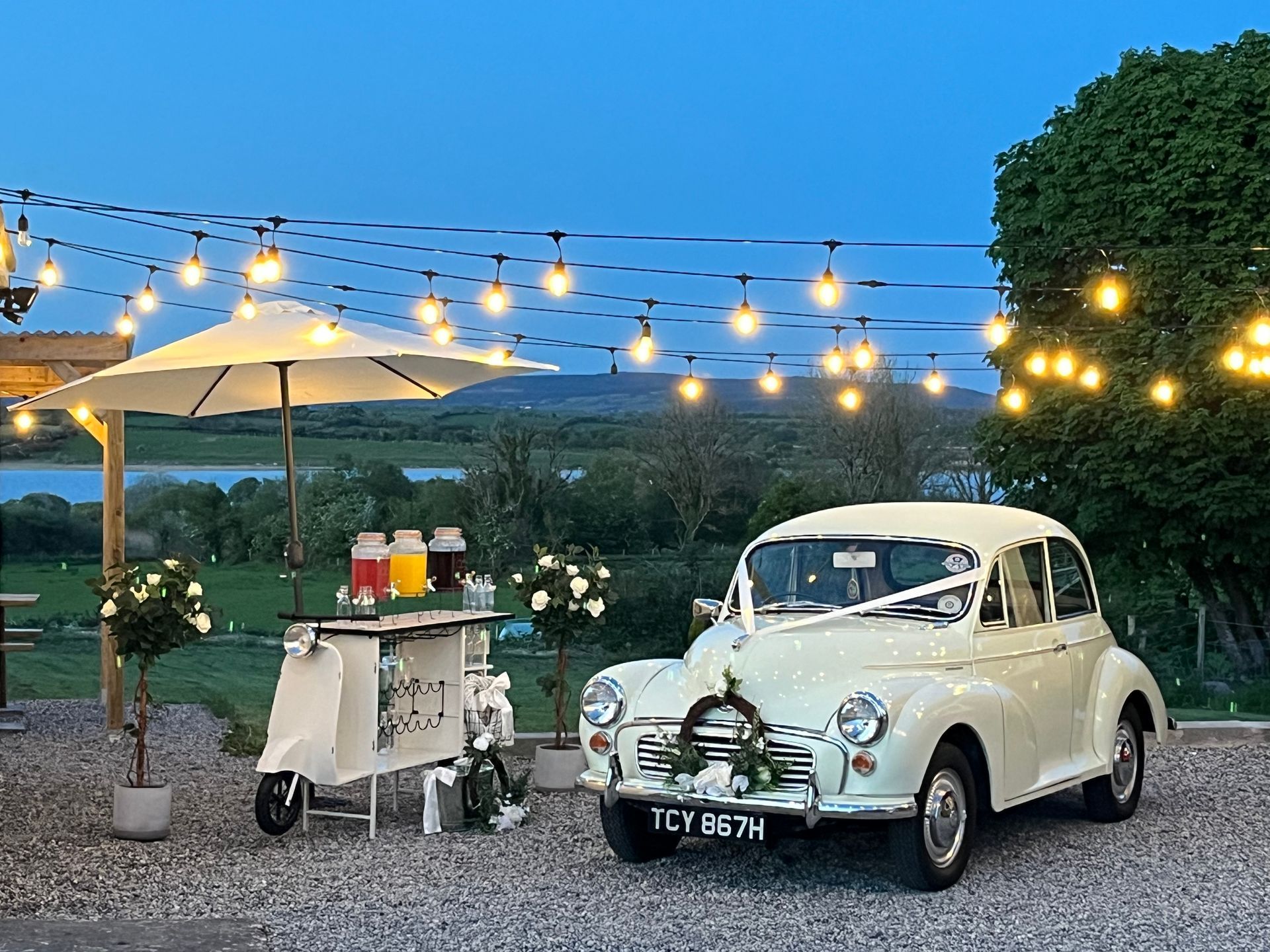 Vintage white car beside a drink cart under string lights at dusk on a lawn