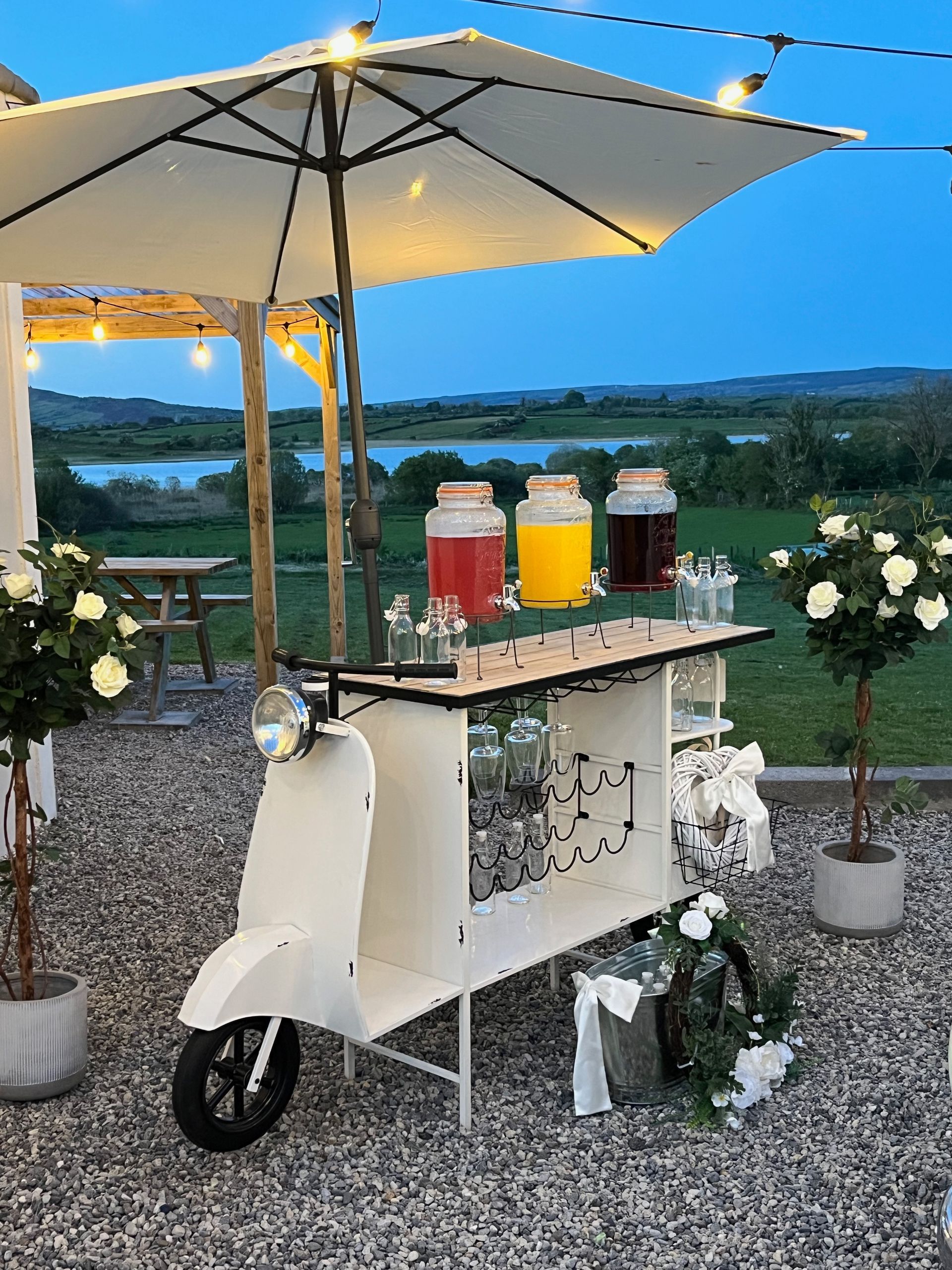 Outdoor drink cart with colorful beverage dispensers under an umbrella at dusk