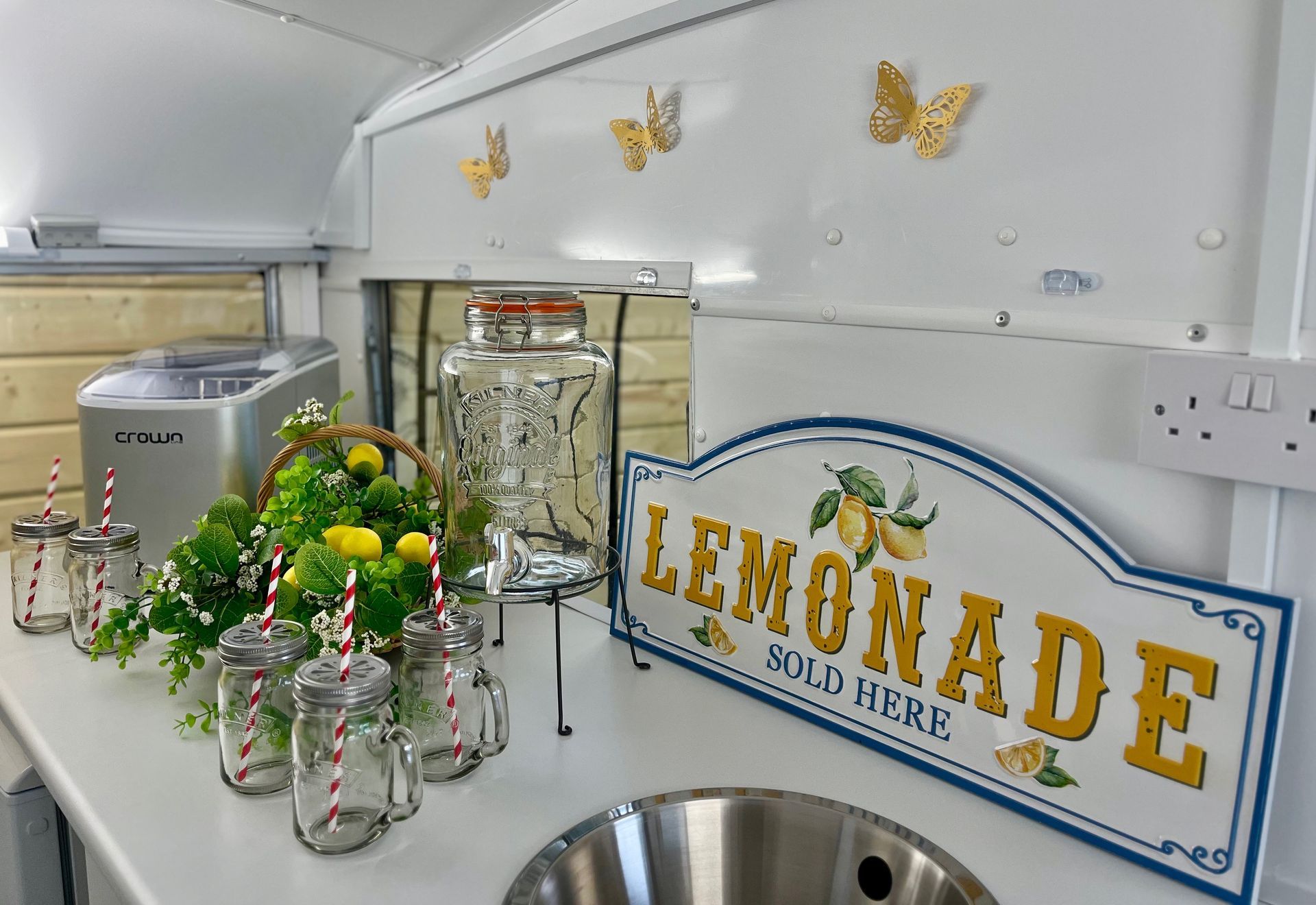 Lemonade stand with jars of lemons, green fruit display, and a “LEMONADE” sign on a countertop.