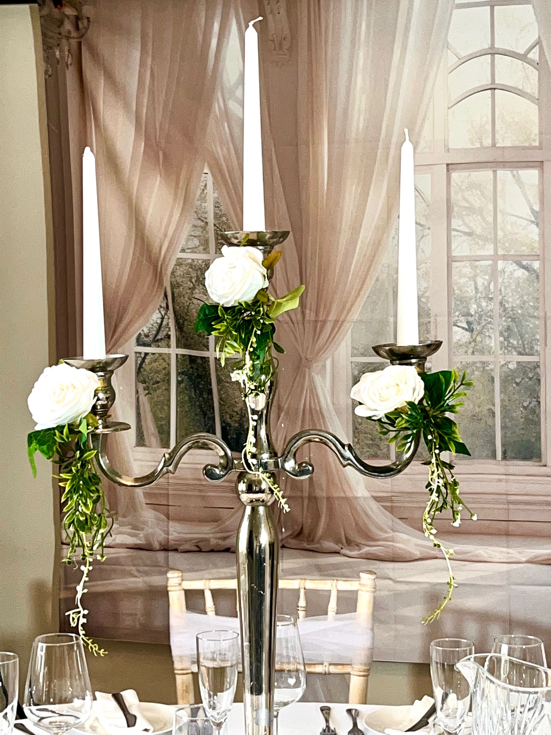 Silver candelabra with white roses and greenery on a set dining table by a curtained window