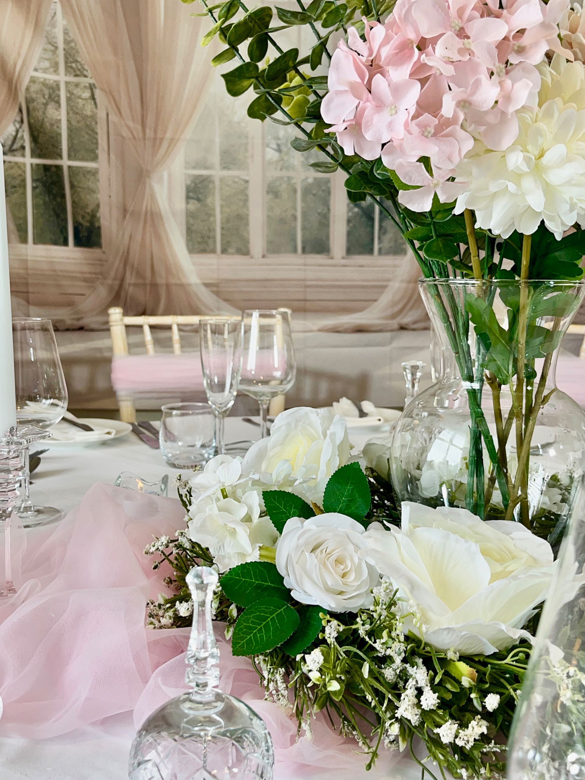 Elegant pastel floral table centerpiece with pink and white flowers, glassware, and soft draped decor