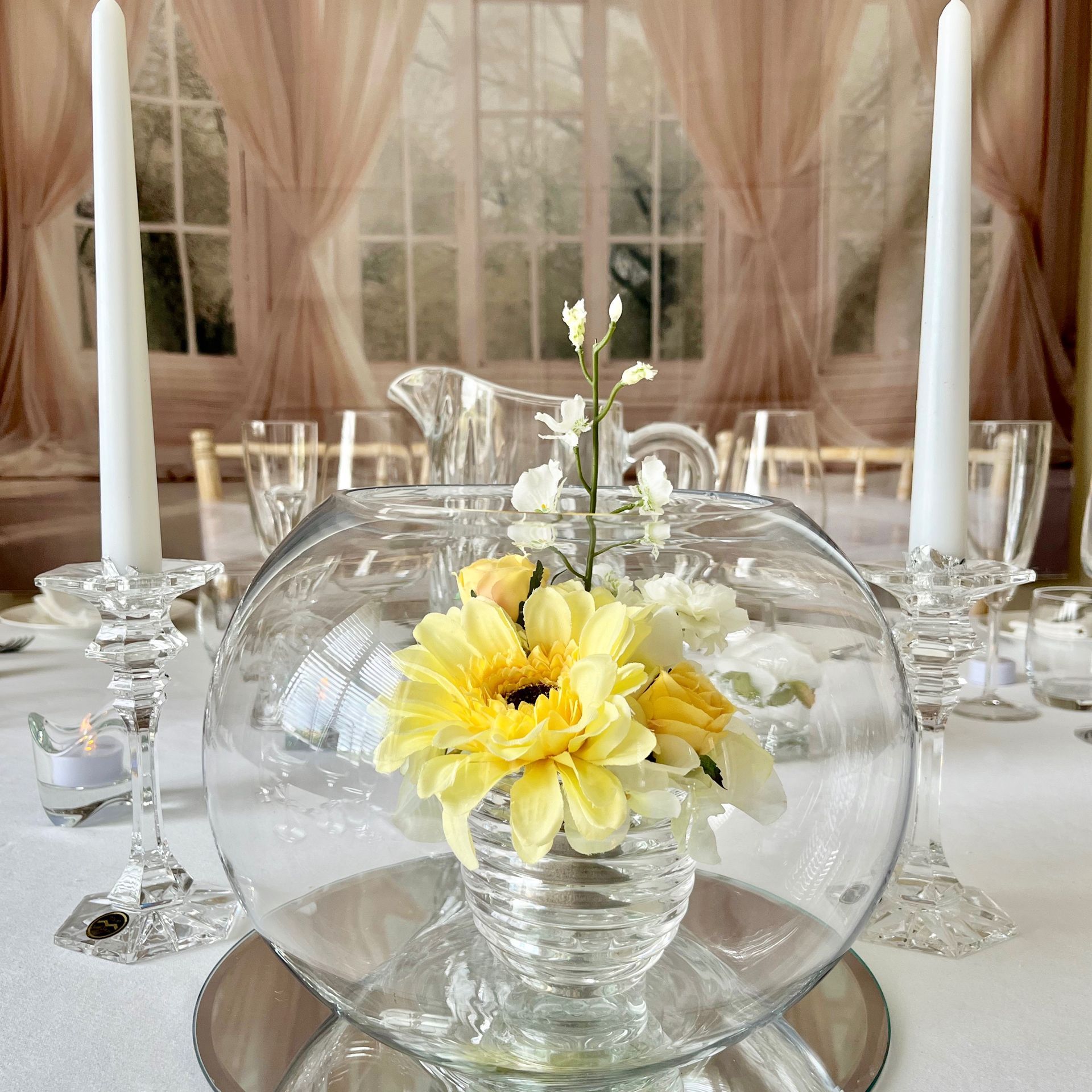 Elegant table setting with a clear glass bowl of yellow flowers and tall white candles by a window