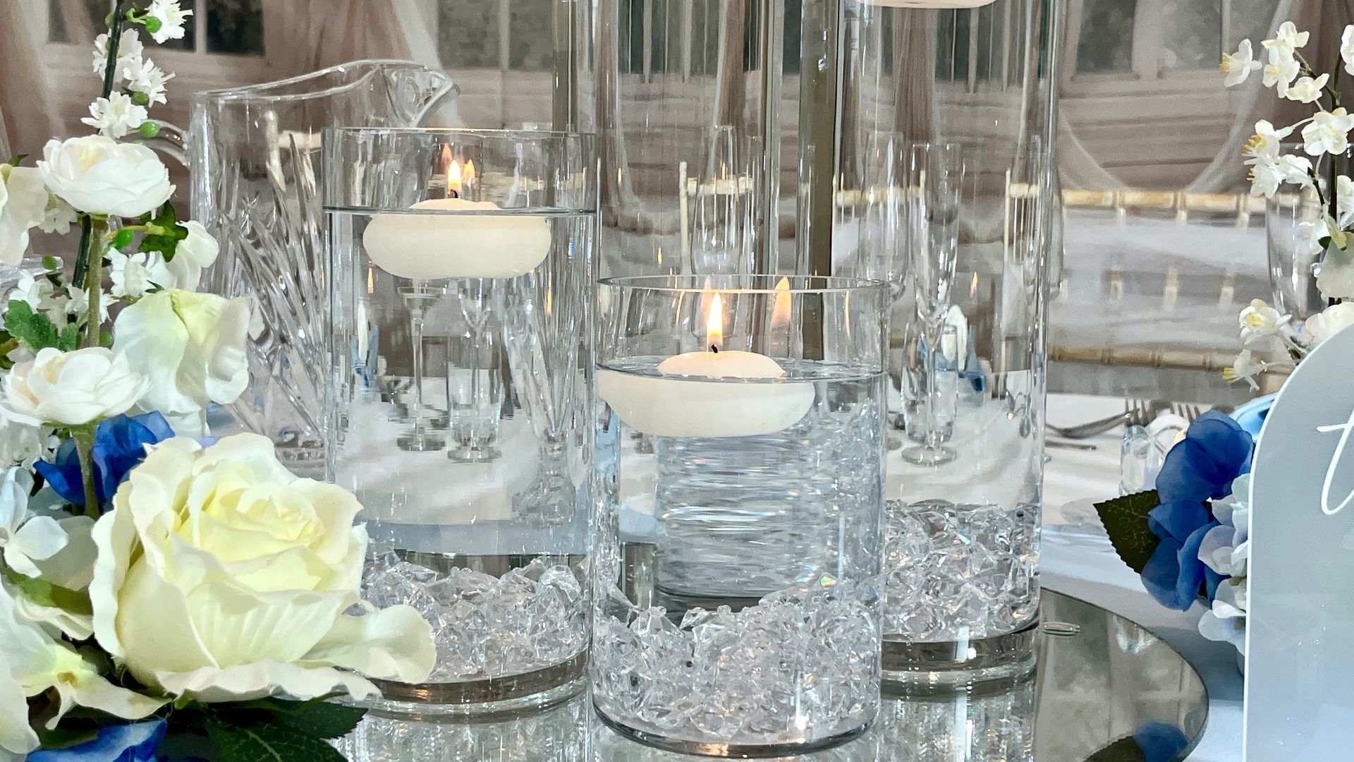Glass candle holders with lit candles and white and blue flowers on a mirrored table