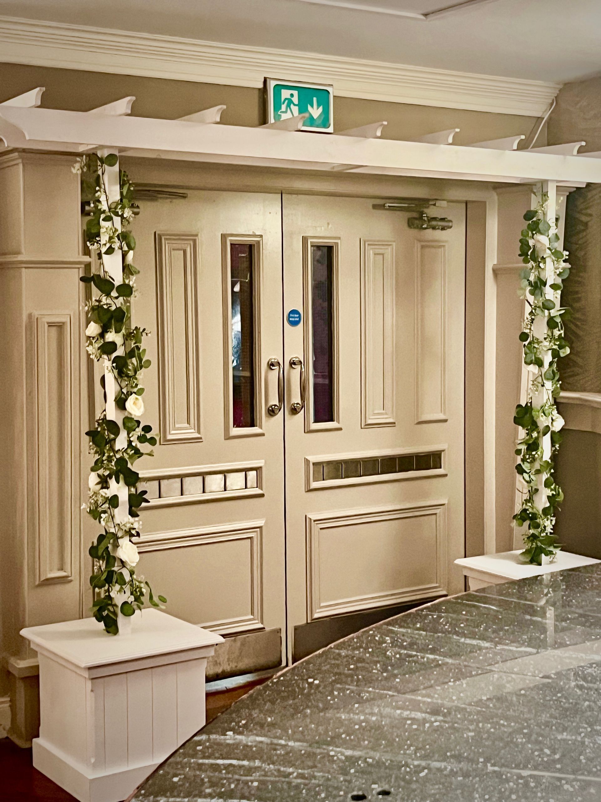 Decorative cream double doors with glass panes, framed by greenery in a carpeted interior hallway