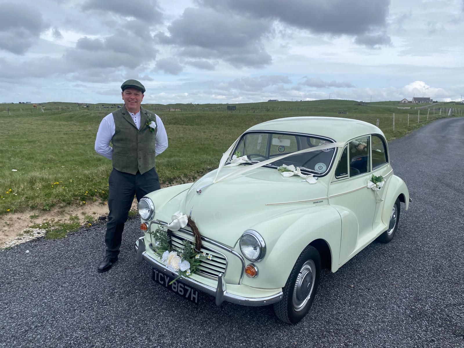 Man standing beside a mint-green vintage car on a gravel road under cloudy skies