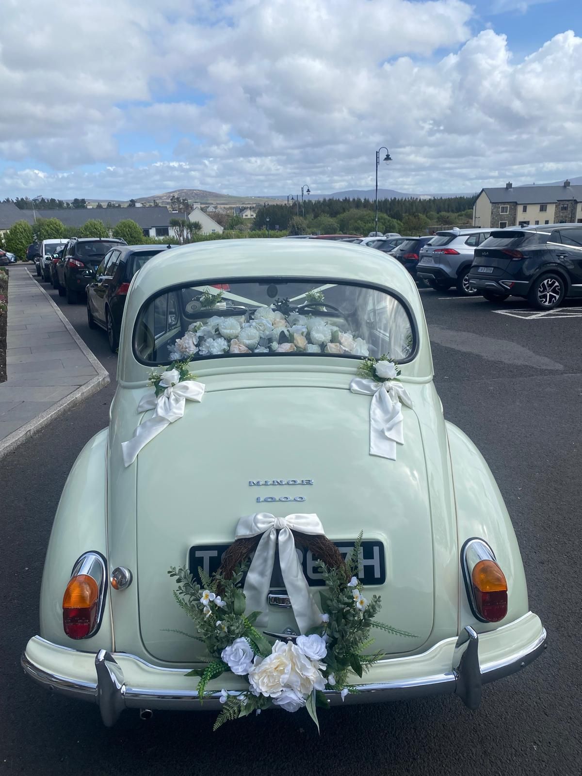 Mint-green vintage car decorated with wedding flowers, parked outdoors under a cloudy sky