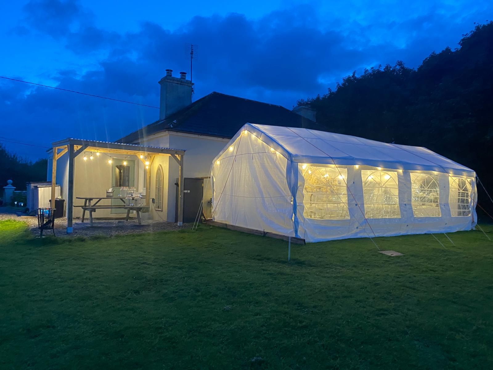 White marquee tent lit with string lights beside a house at dusk on a grassy lawn