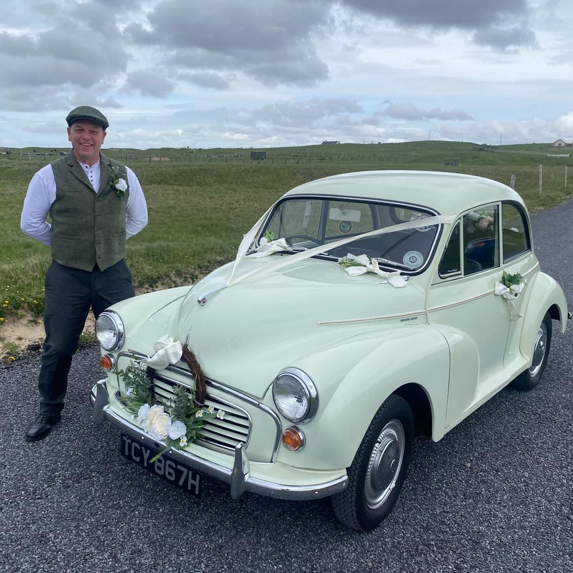 Man standing beside a pale green vintage car on a rural road under cloudy skies